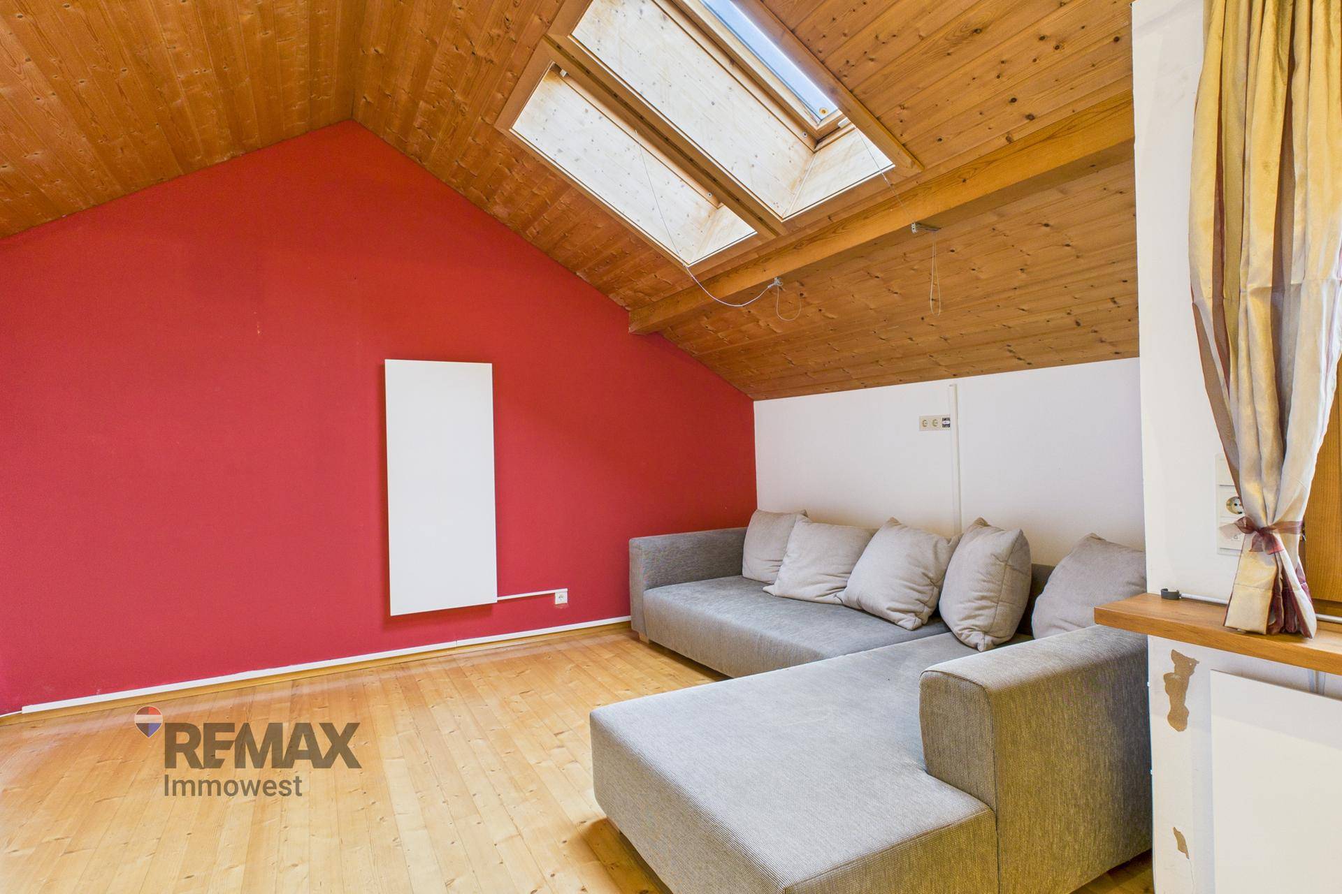 Gemütliches Wohnzimmer mit L-förmigem Sofa, roter Wand und Holzboden unter Dachschrägenfenstern.