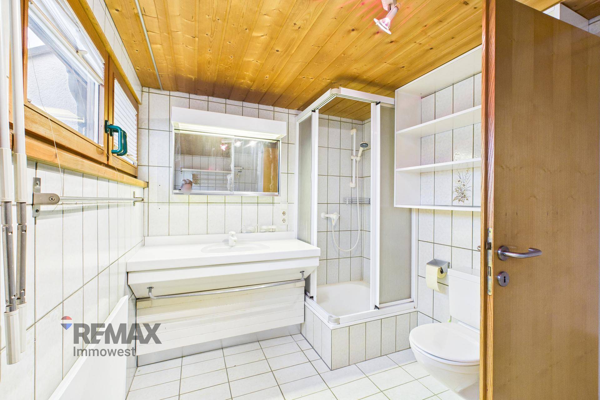 Gefliestes Badezimmer mit Duschkabine, Einzelwaschbecken und Fenster, unter einer Holzdecke.