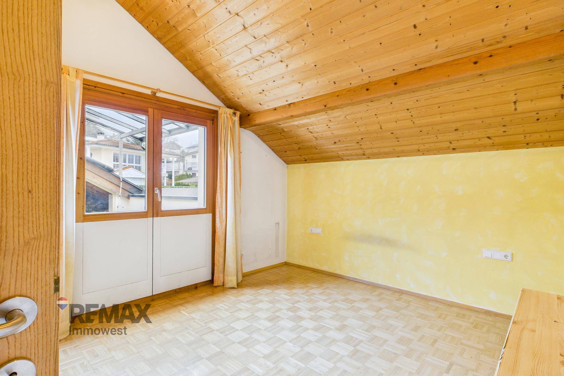 Schlafzimmer mit Parkettboden, gelber Wand und Zugang zu einem Außenbereich, unter einer Holzdecke.