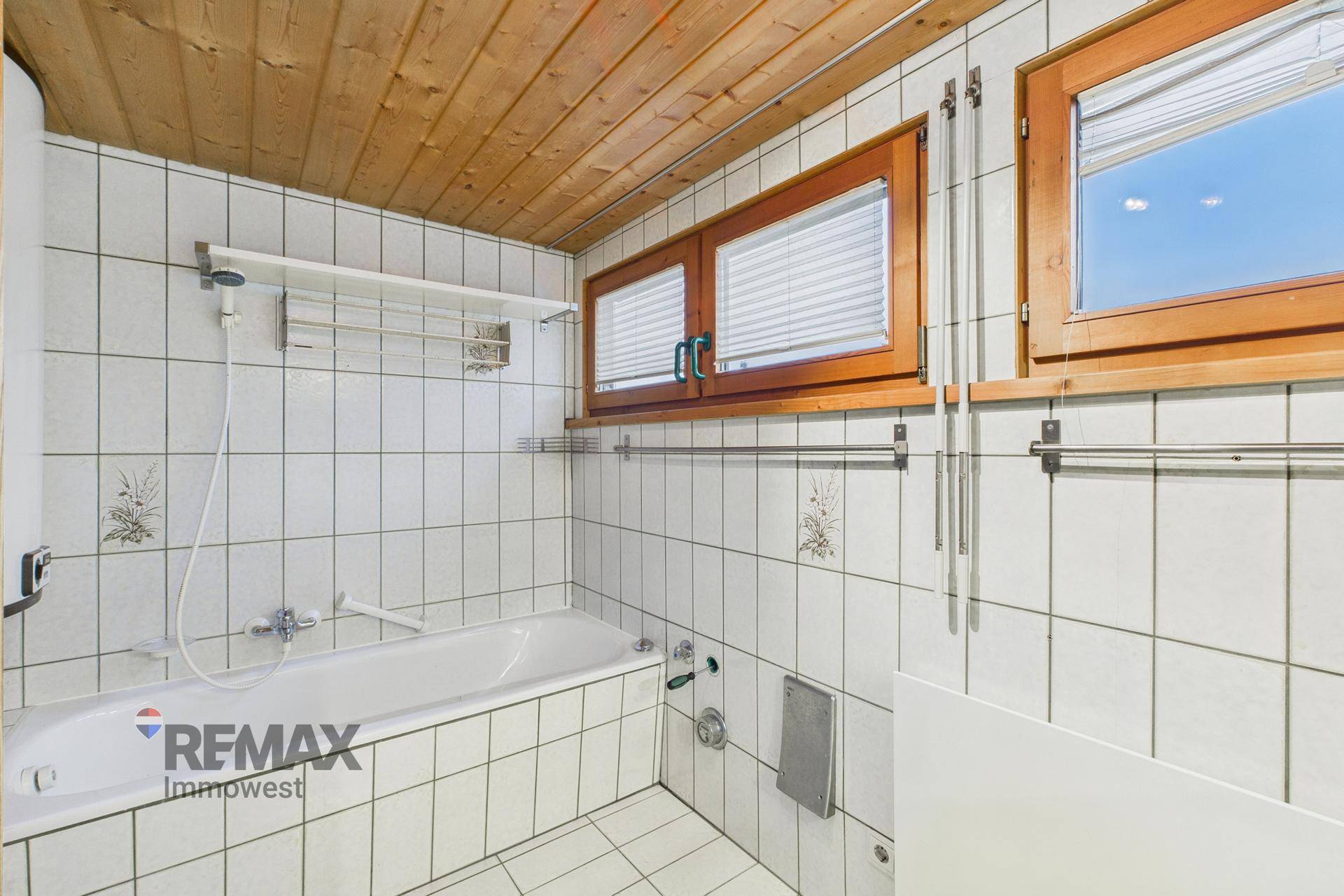 Badezimmer mit Badewanne, weißen Fliesen und Holzdecke, gut belichtet durch zwei Fenster.