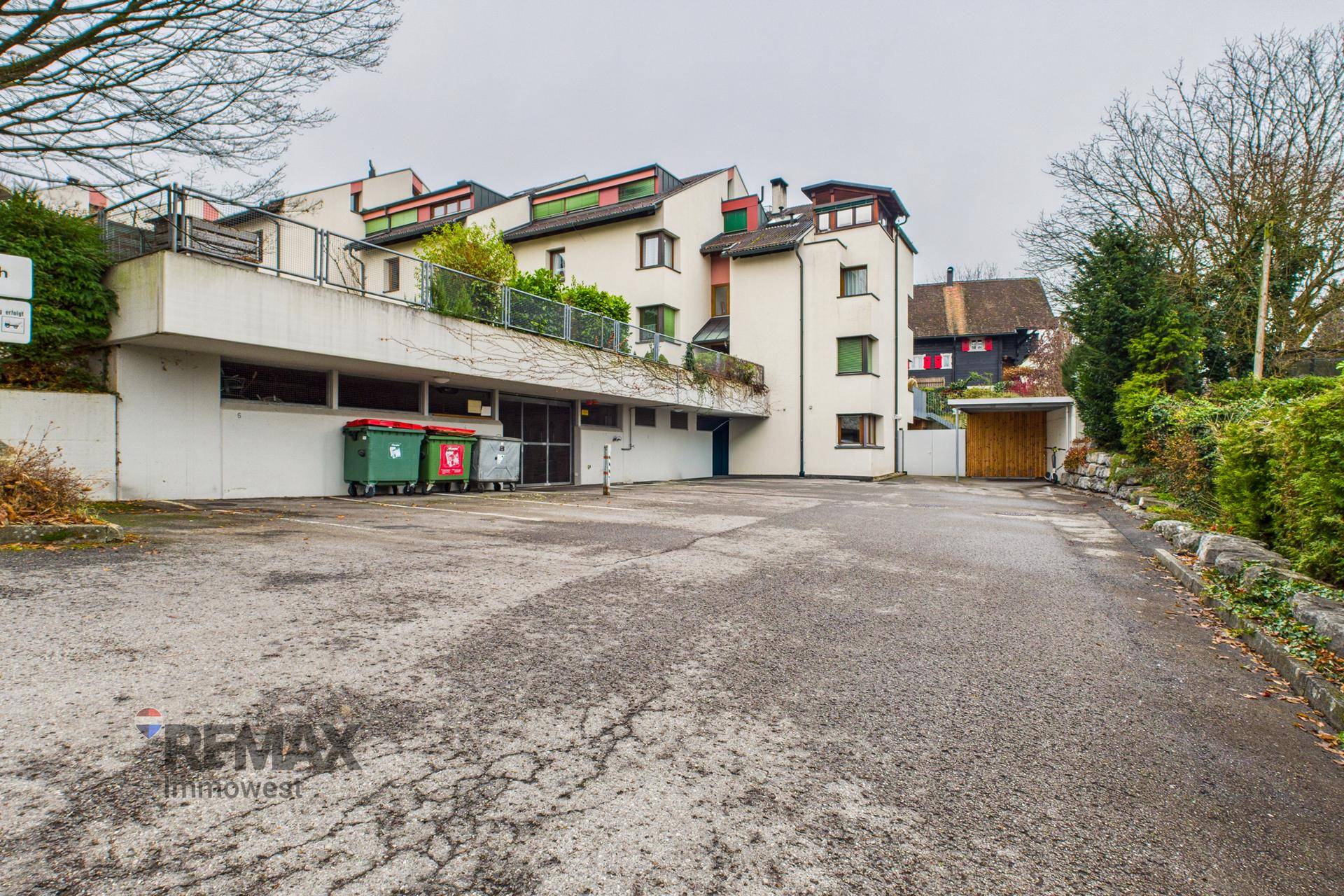 Außenansicht der Wohnanlage mit Parkplatz und Zufahrt zur Tiefgarage, umgeben von Bäumen.