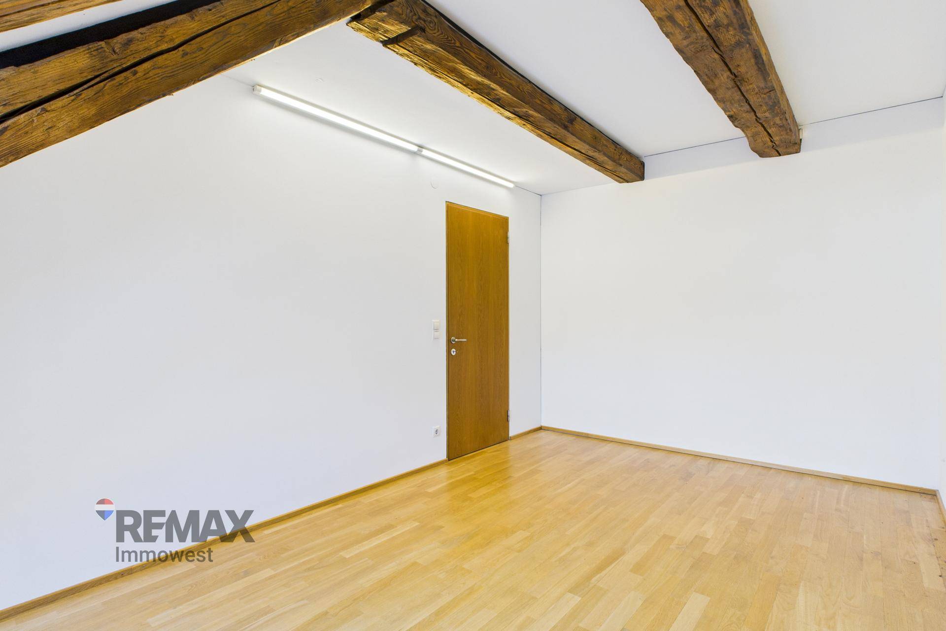 Leeres Zimmer mit Holzbalken an der Decke und hellem Parkettboden.