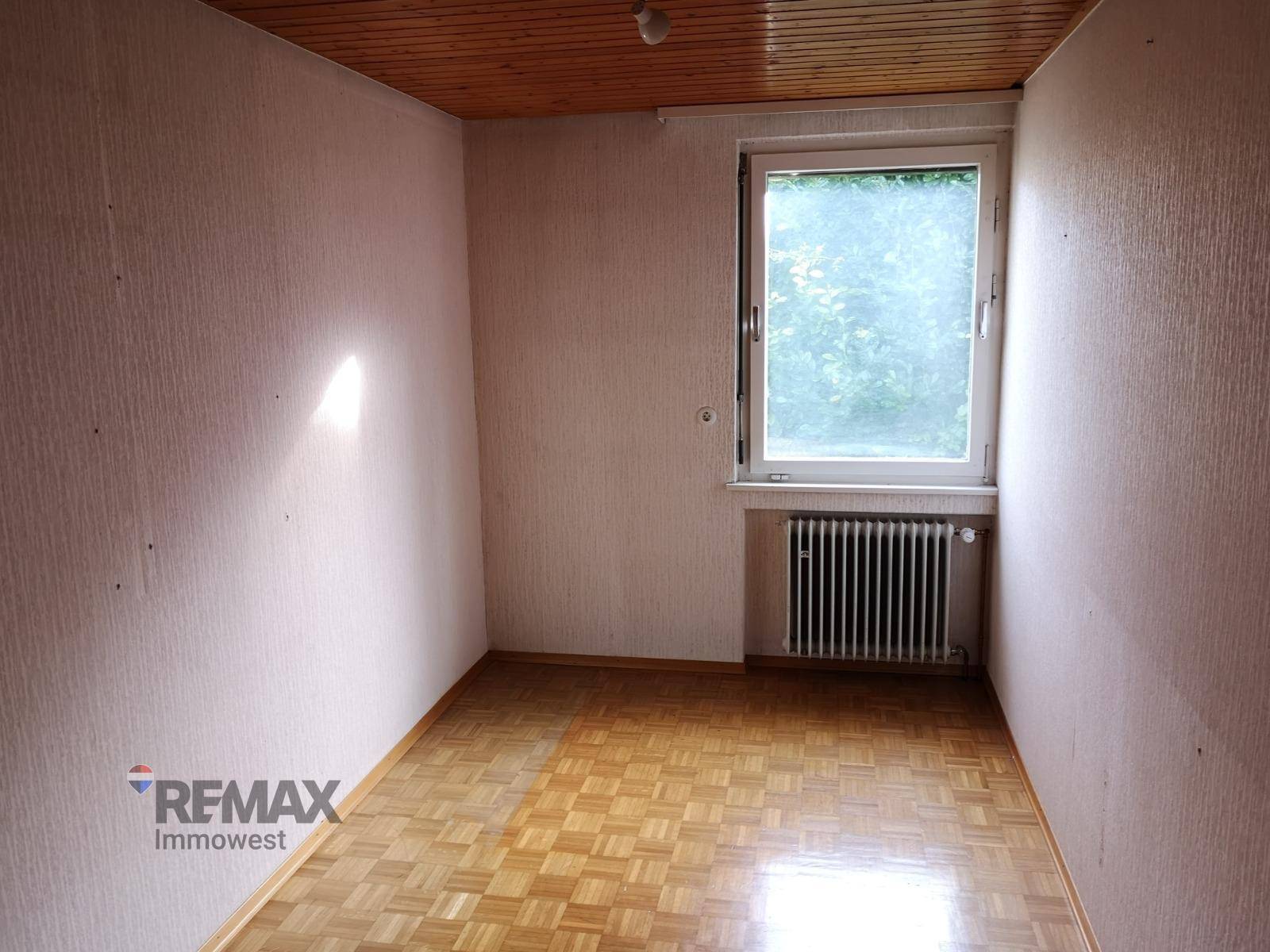 Leeres Zimmer mit Parkettboden, Holzdecke und Fenster mit Blick ins Grüne.