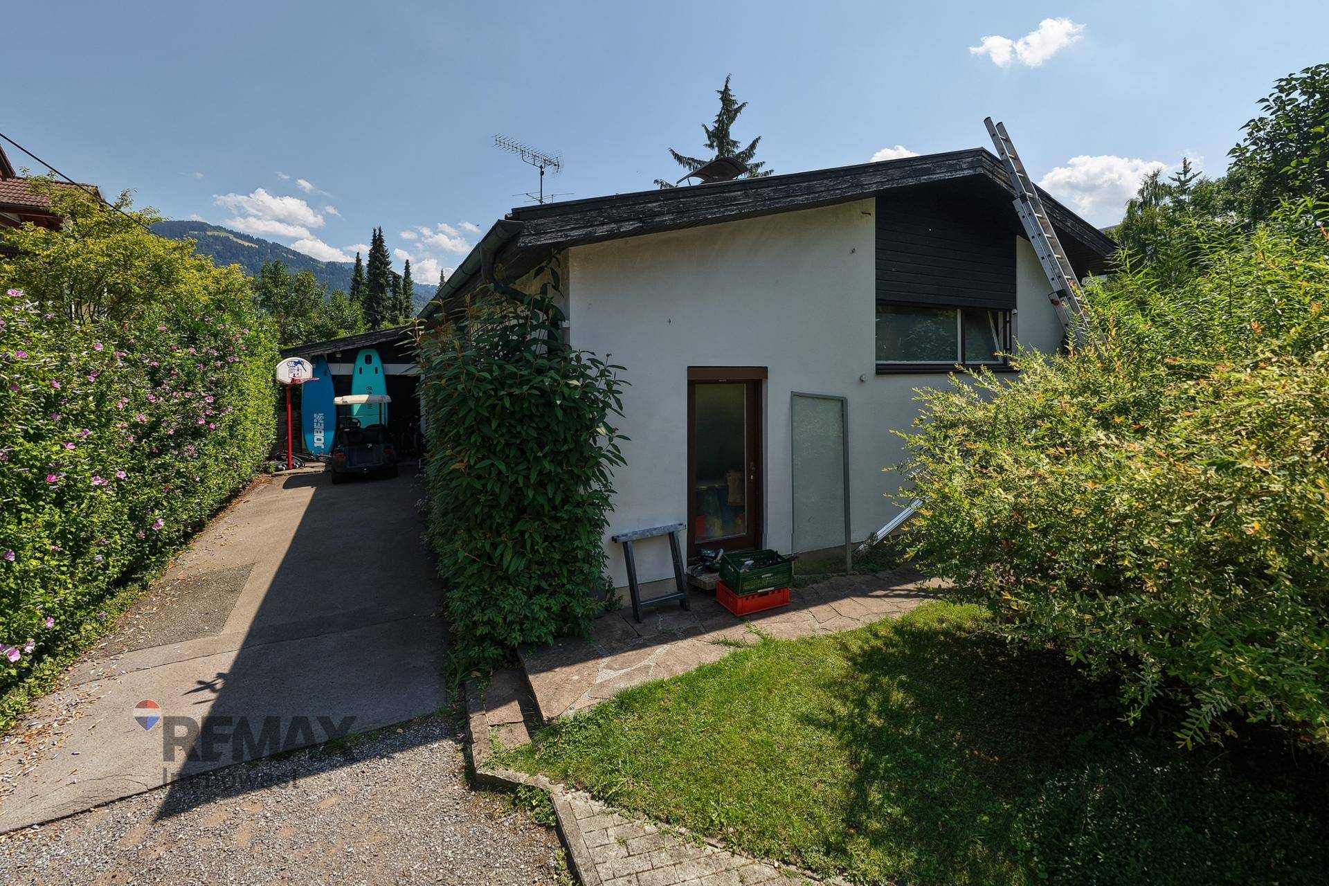 Seitenansicht des Hauses mit Einfahrt, Hecke und Blick auf die Berge im Hintergrund.