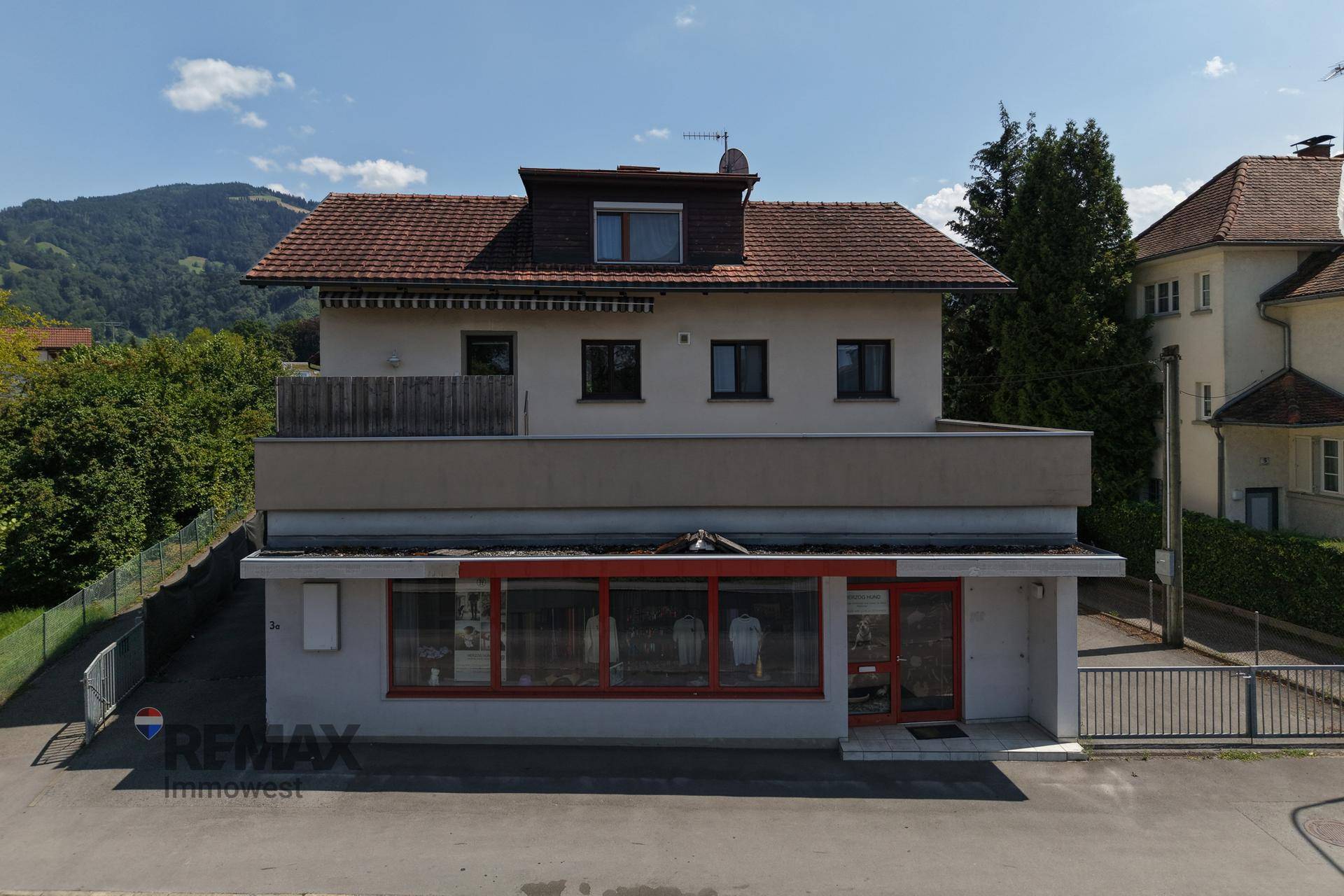 Gebäude mit Geschäftsfläche im Erdgeschoss und Wohnungen darüber, umgeben von Grün.