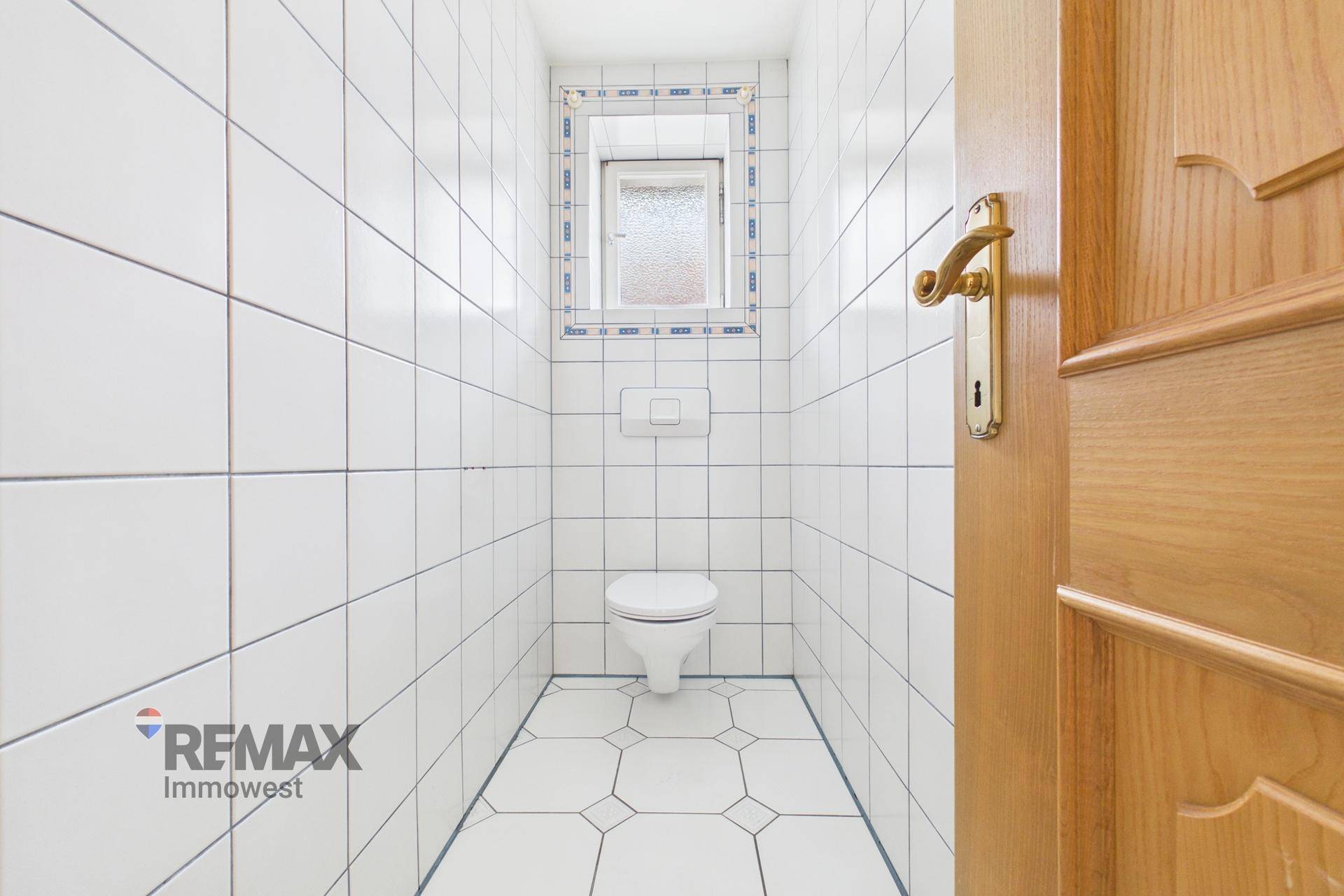 Kleines WC mit weißen Fliesen, Toilette und Fenster, das Tageslicht hereinlässt.