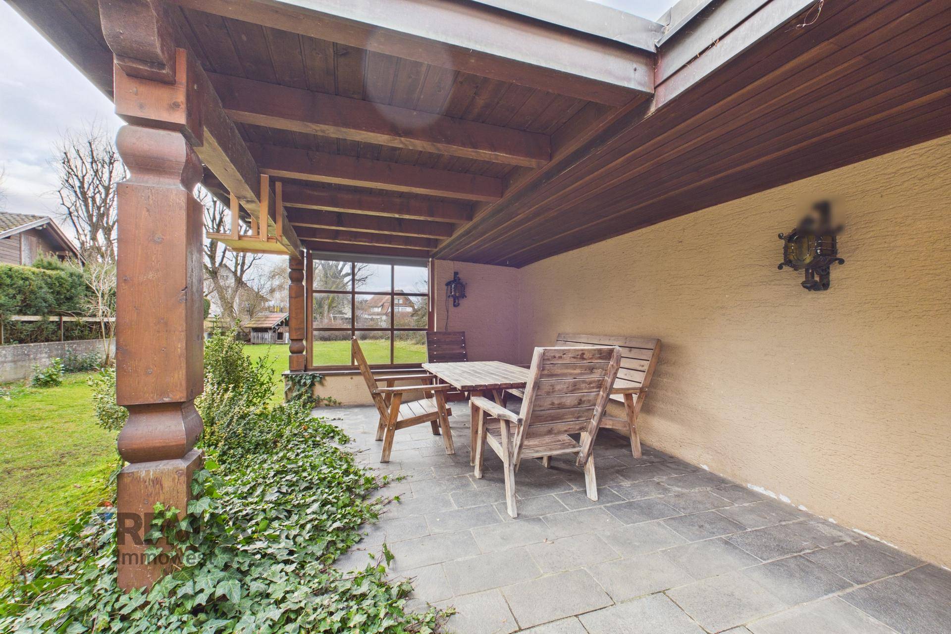 Überdachte Terrasse mit Holztisch und Stühlen, umgeben von Grün und Blick in den Garten.