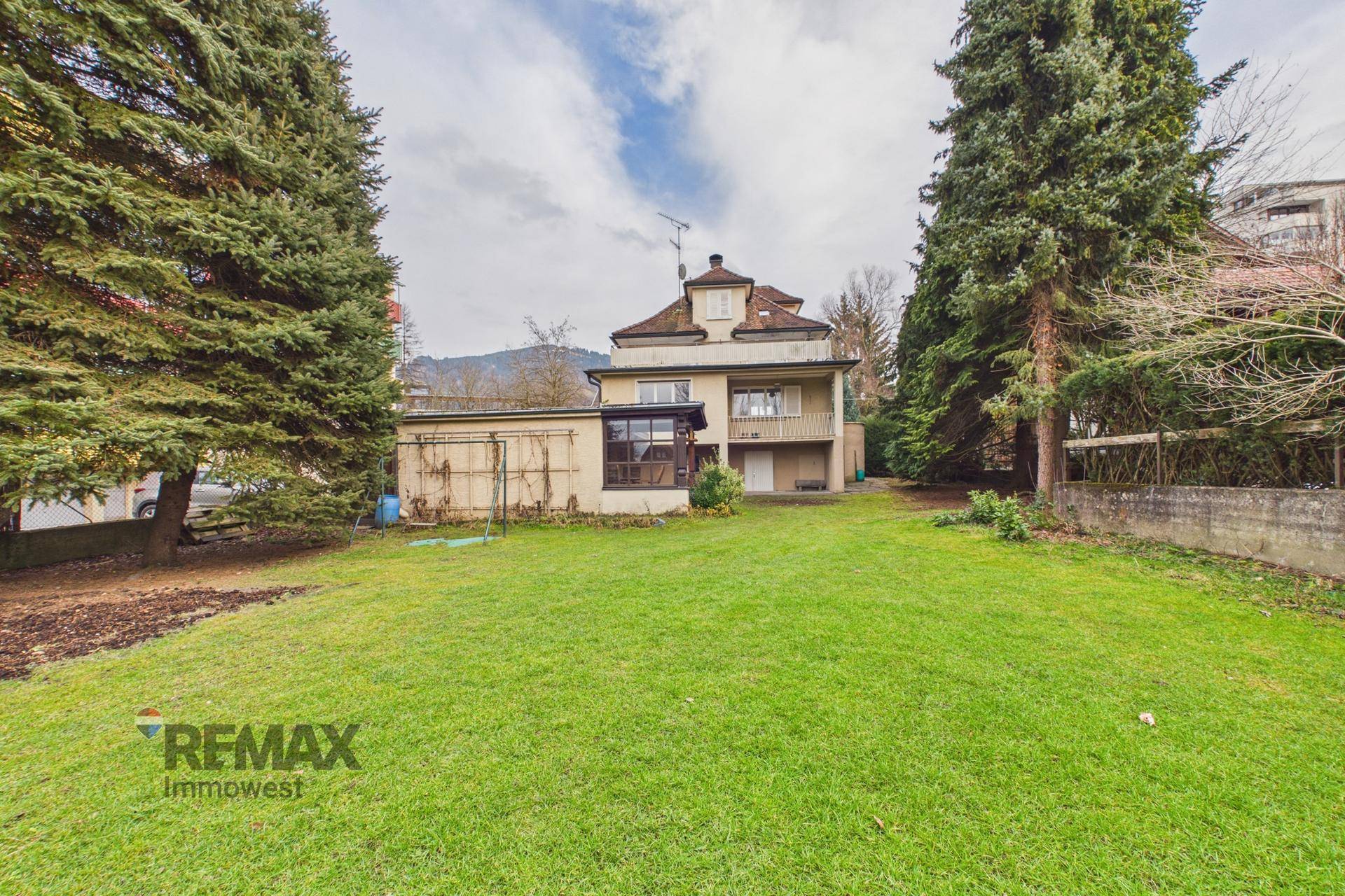 Ansicht des Hauses von der weitläufigen Gartenfläche mit hohen Bäumen und Bergen im Hintergrund.