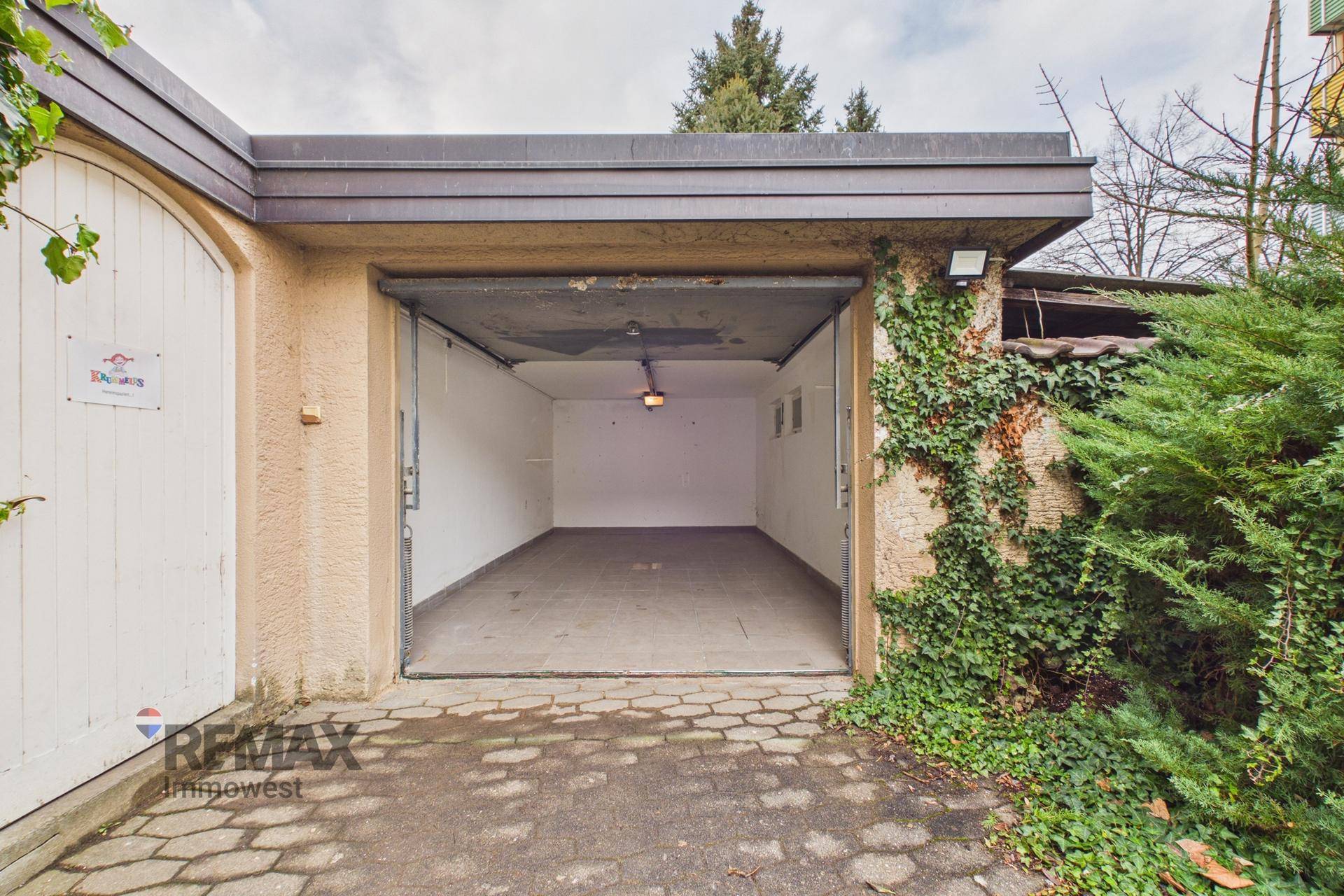Leere Garage mit offenem Tor und gepflastertem Vorplatz, umgeben von Efeu.
