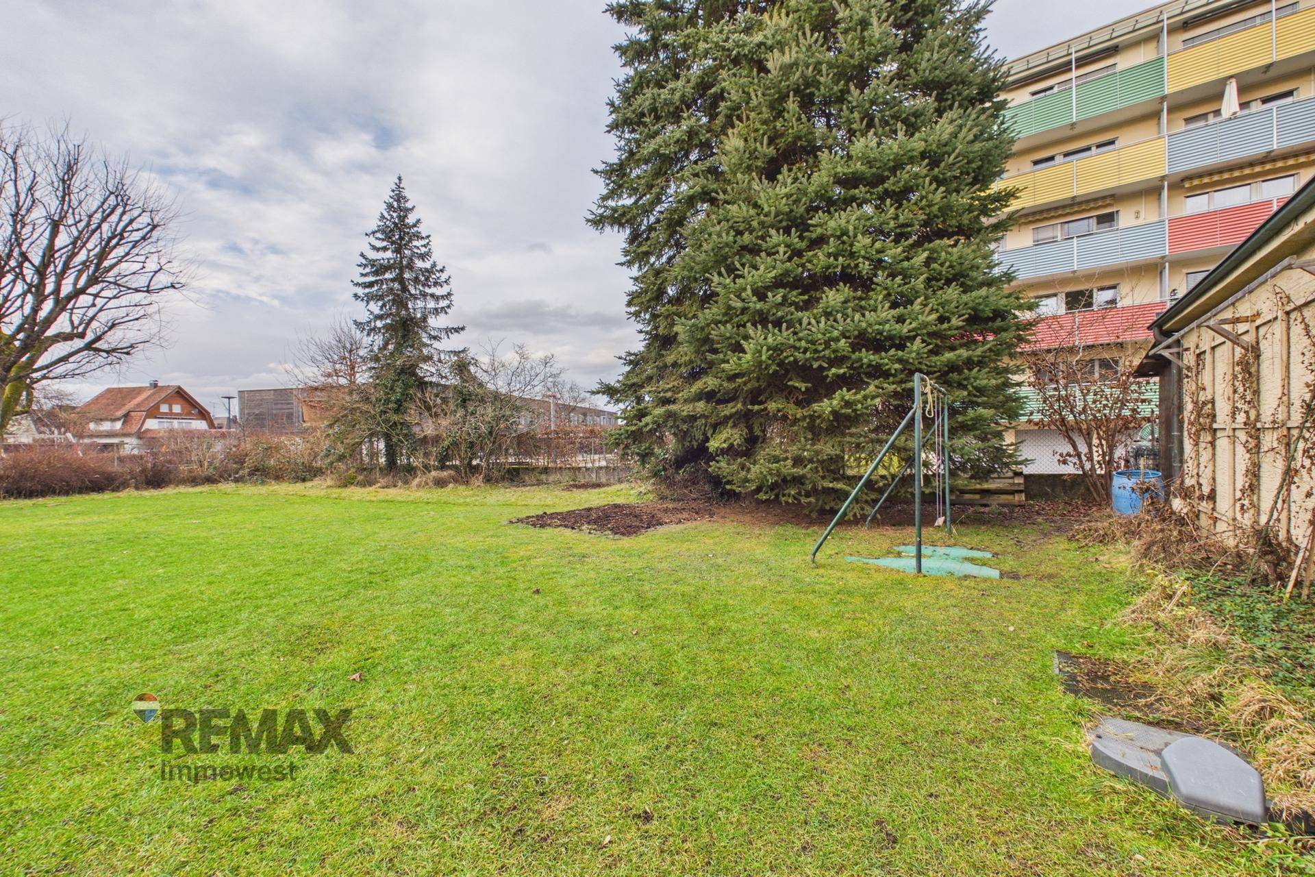 Großer Garten mit Schaukel und hohen Bäumen, angrenzend an ein Mehrfamilienhaus.