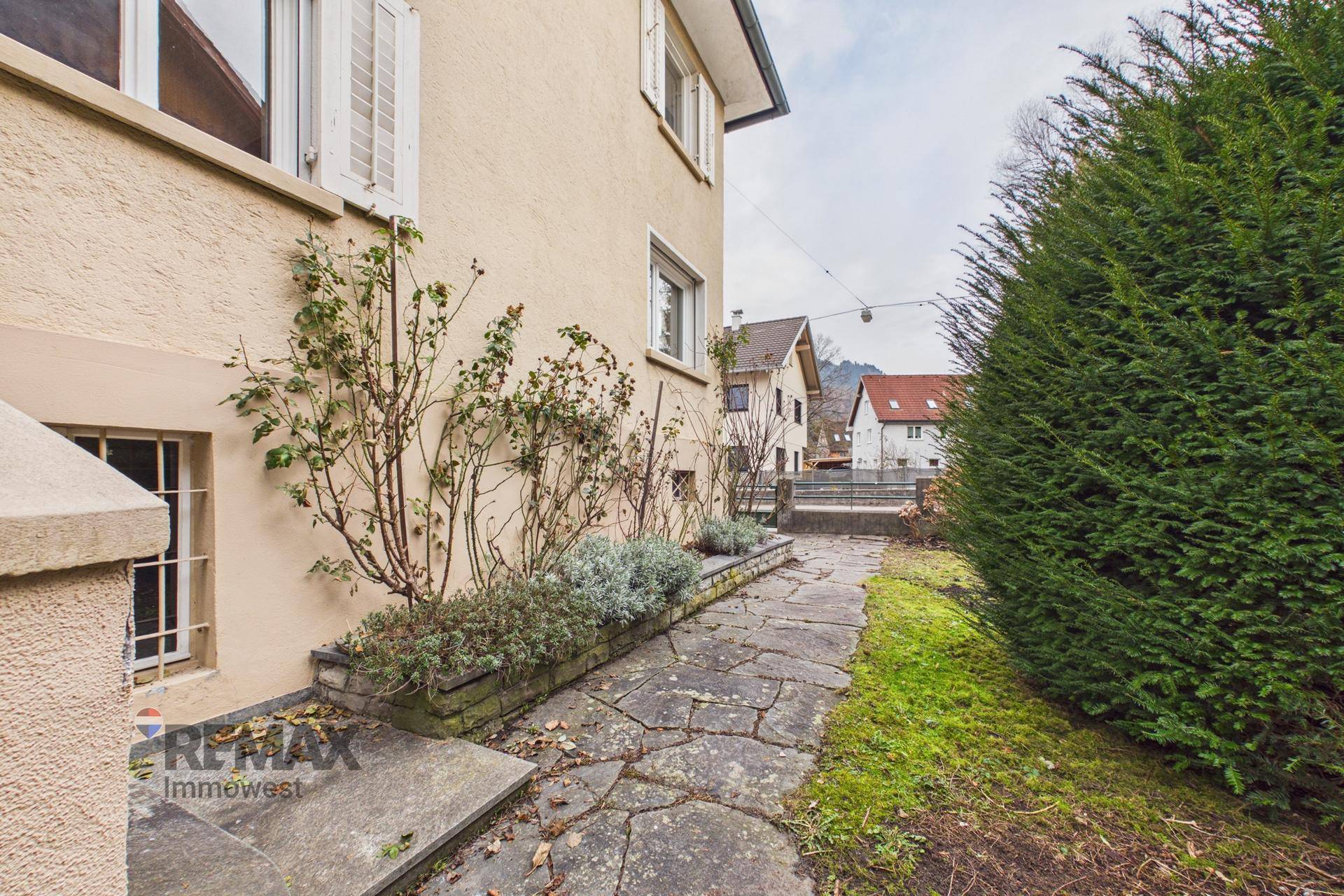 Seitenansicht des Hauses mit Steinweg und bepflanzten Beeten entlang der Fassade.