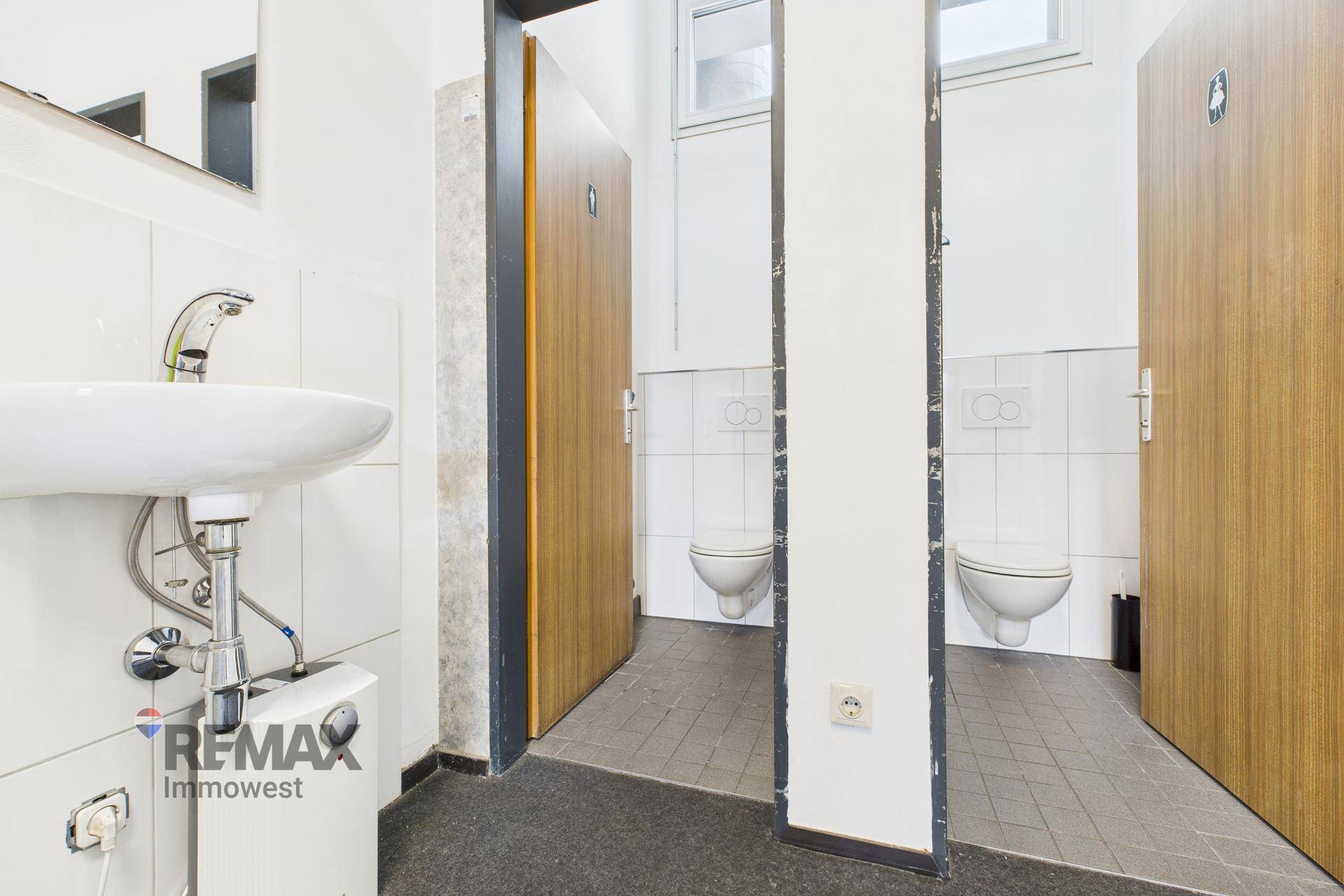 Toilettenraum mit Waschbecken, Wand-WCs und weißen Fliesen an den Wänden.