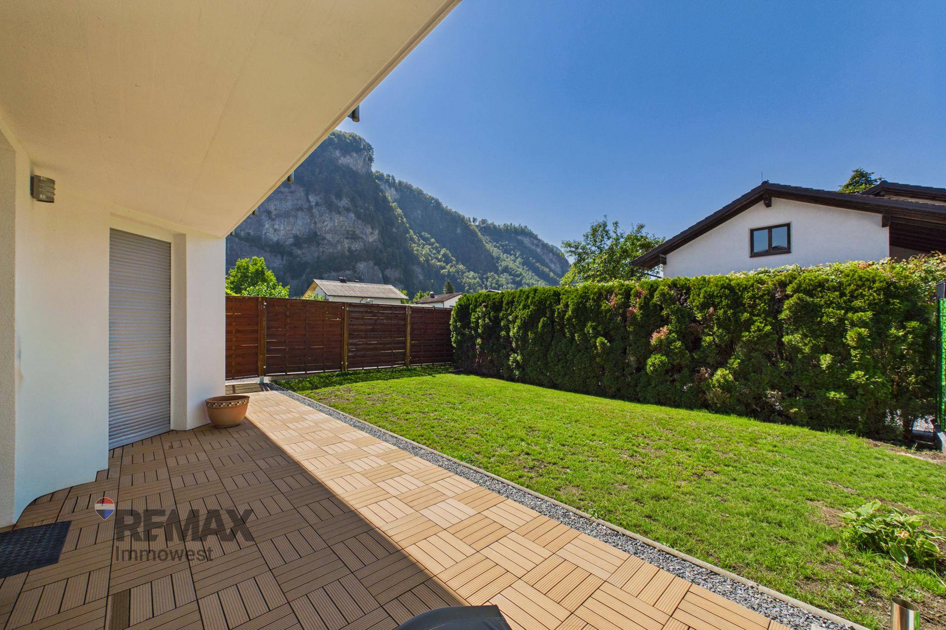 Sonniger Gartenbereich mit Holzterrasse, Rasenfläche und Blick auf die umliegenden Berge.