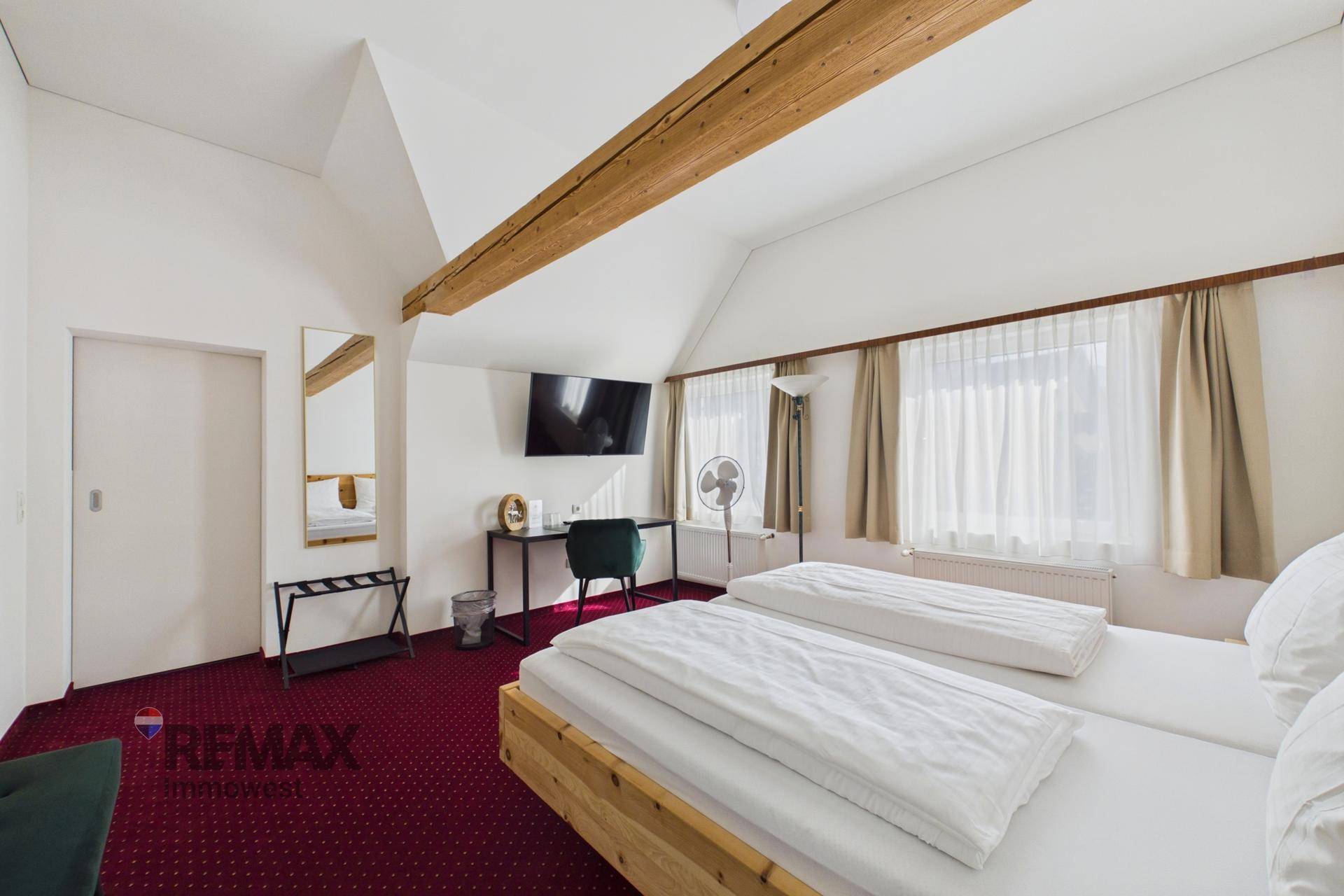 Dachgeschoss-Schlafzimmer mit sichtbaren Holzbalken, Doppelbett und rotem Teppichboden, bietet rustikalen Charme.
