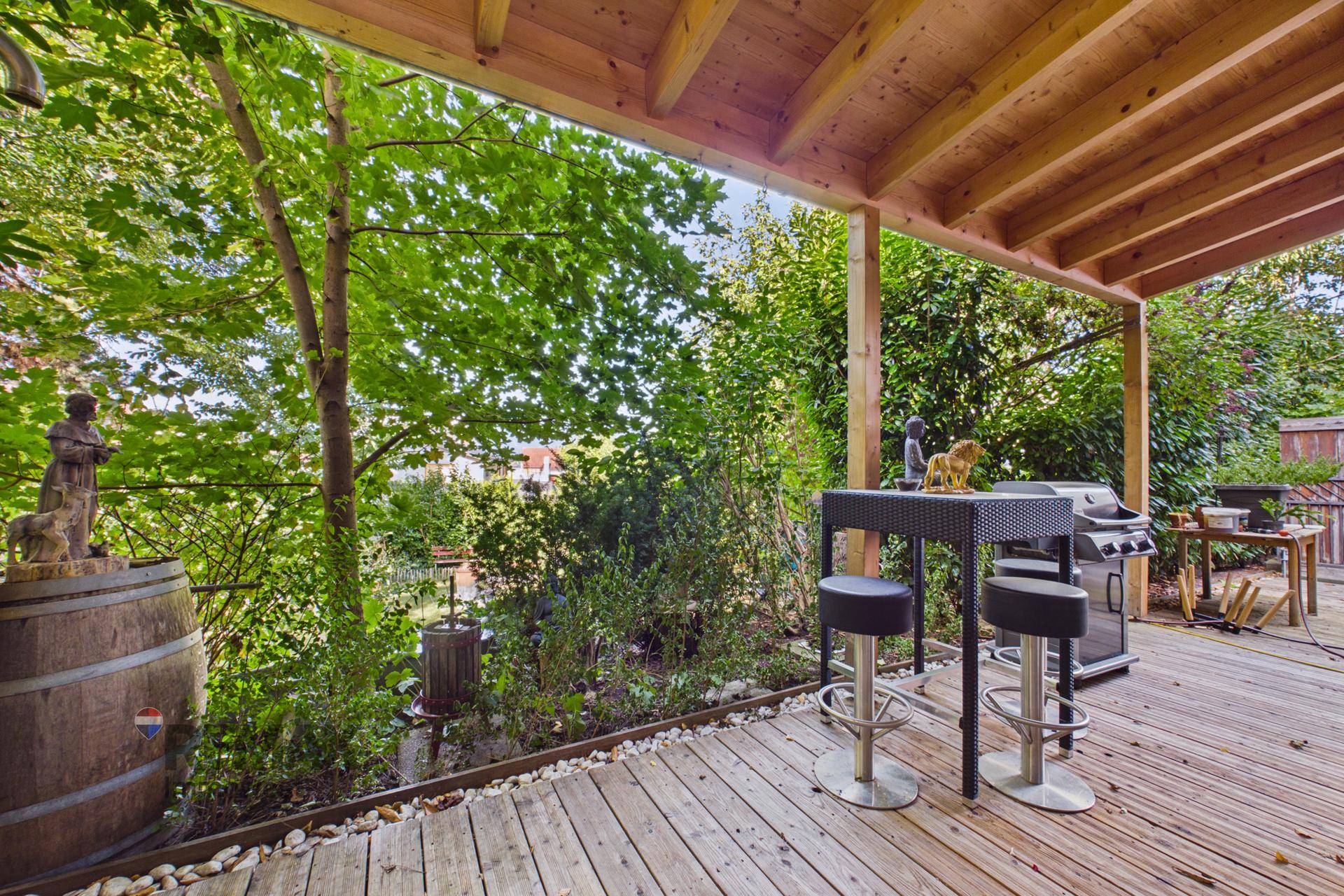 Überdachte Holzterrasse mit Bartisch, Hockern und Grillbereich, umgeben von üppigem Grün und Bäumen.