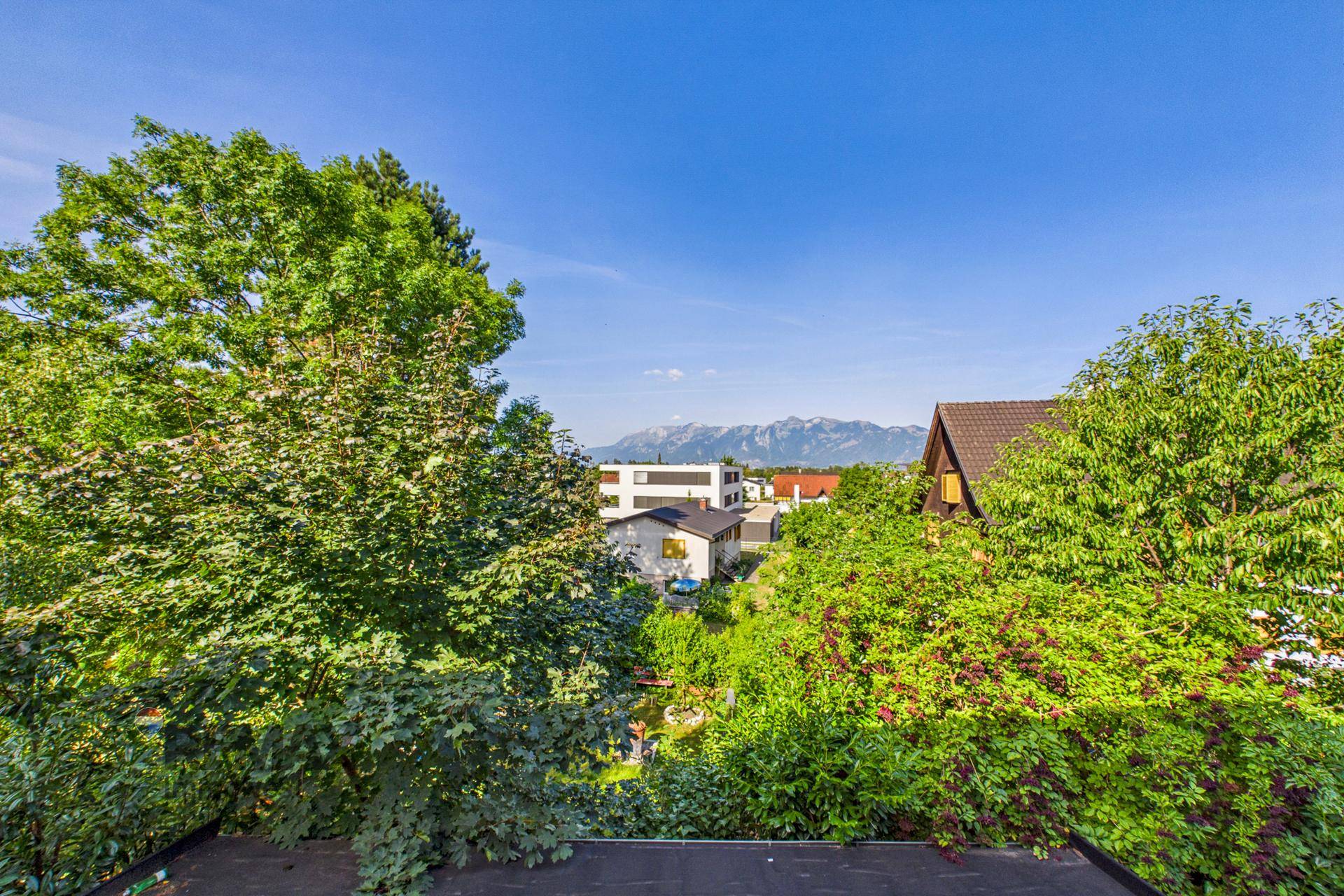 Weitblick über grüne Bäume und Dächer von Häusern mit majestätischen Bergen im Hintergrund unter blauem Himmel.