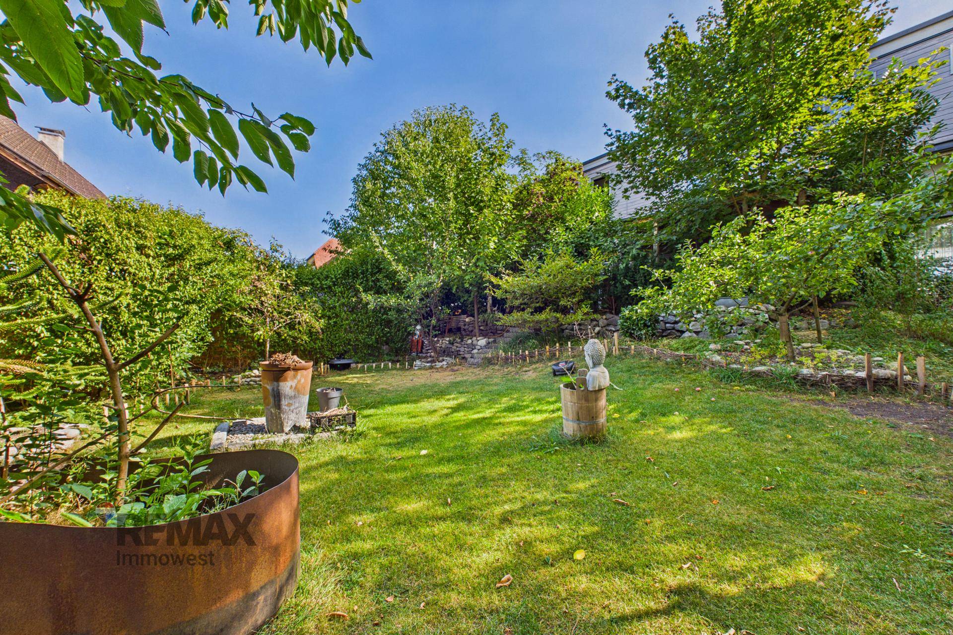 Gepflegter Gartenbereich mit Rasenfläche, Bäumen und dekorativen Elementen unter strahlendem Sonnenschein.