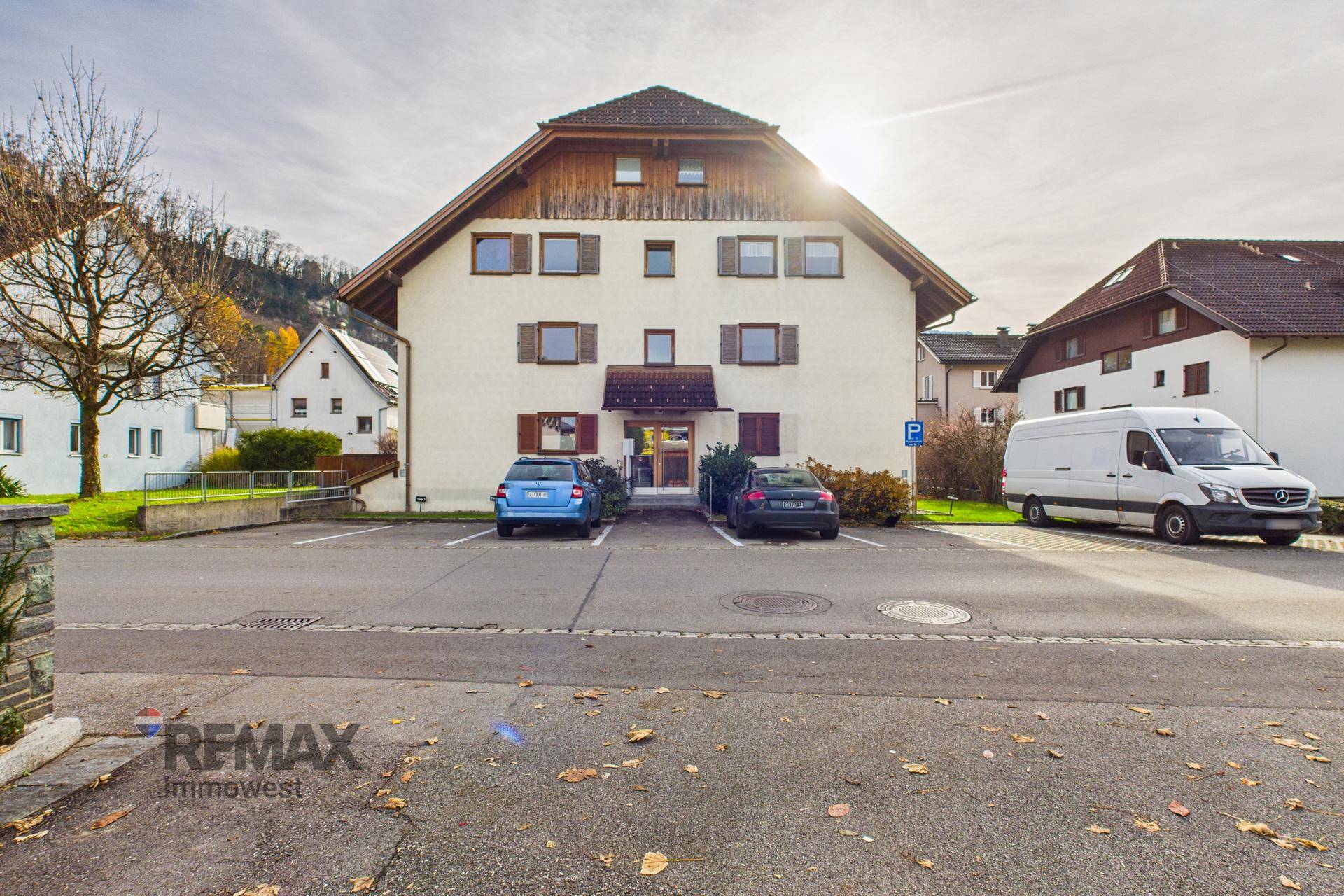 Vorderansicht eines Mehrfamilienhauses mit Parkplätzen und umliegender Natur.