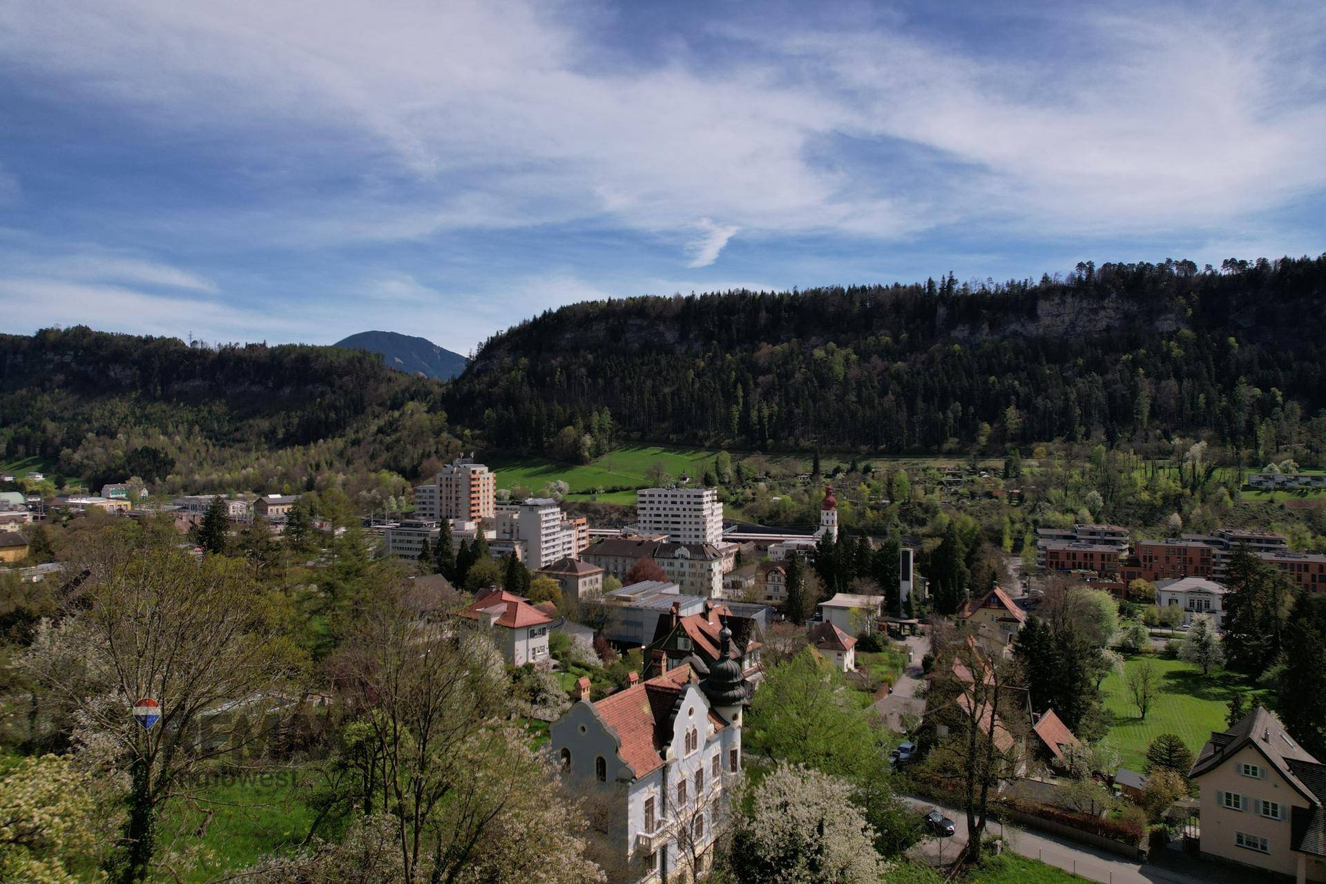 Weitläufiger Blick über eine grüne Stadtlandschaft mit bewaldeten Hügeln und einem historischen Gebäude im Vordergrund.