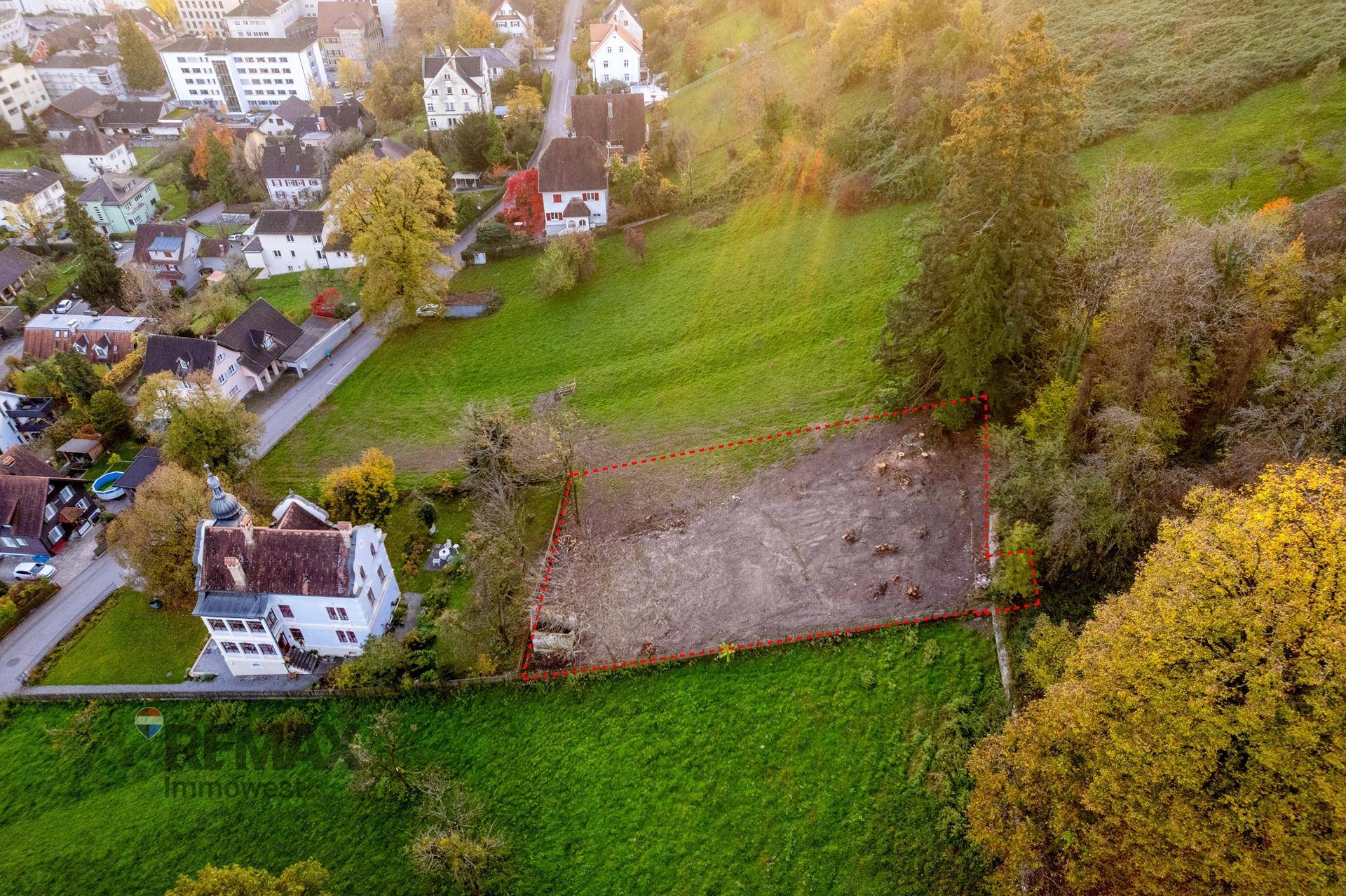Luftaufnahme eines Baugrundstücks, rot markiert, neben einem Gebäude und umgeben von grüner Landschaft.