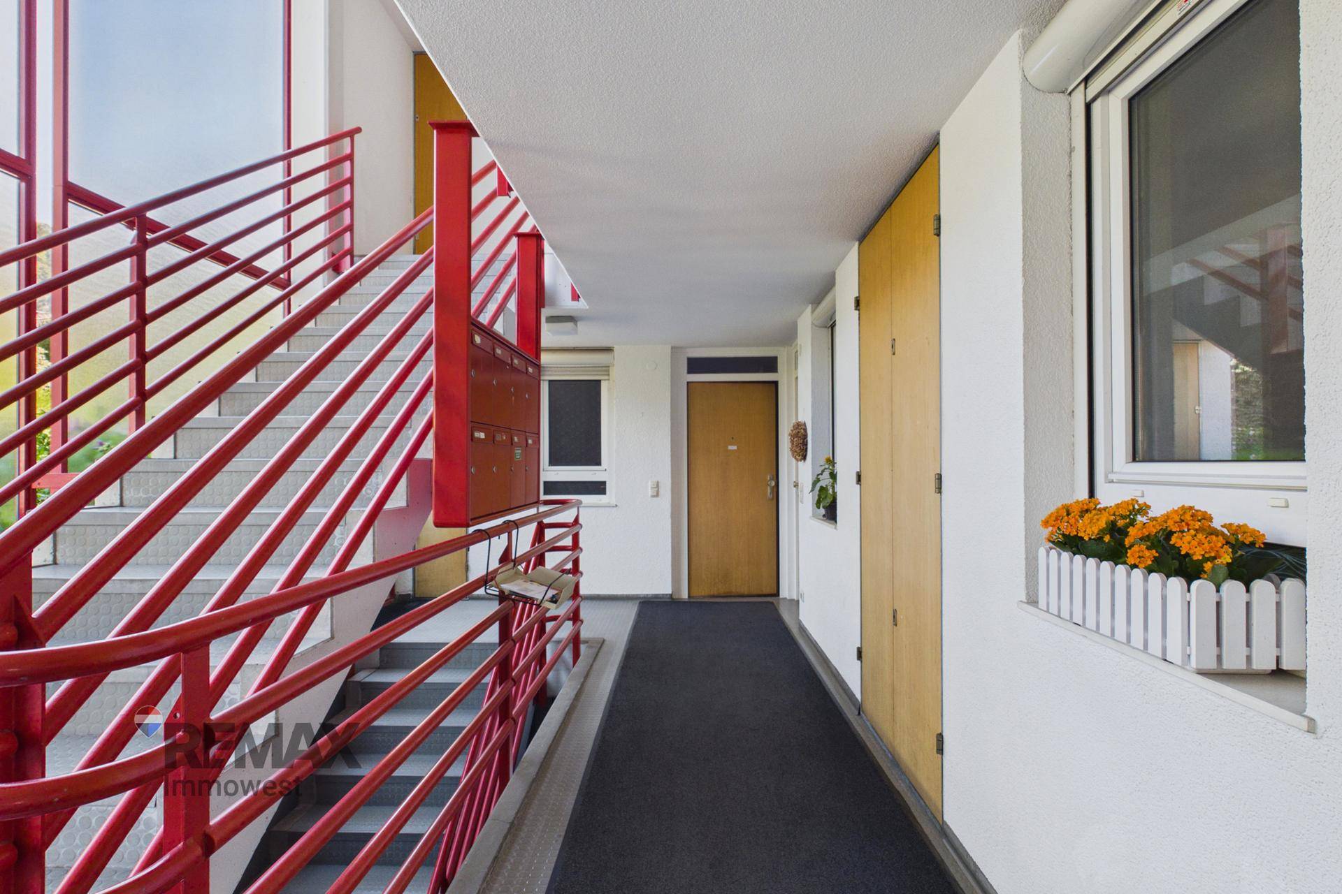 Heller Treppenaufgang und Flur im Mehrfamilienhaus mit roten Geländern und Fenstern.