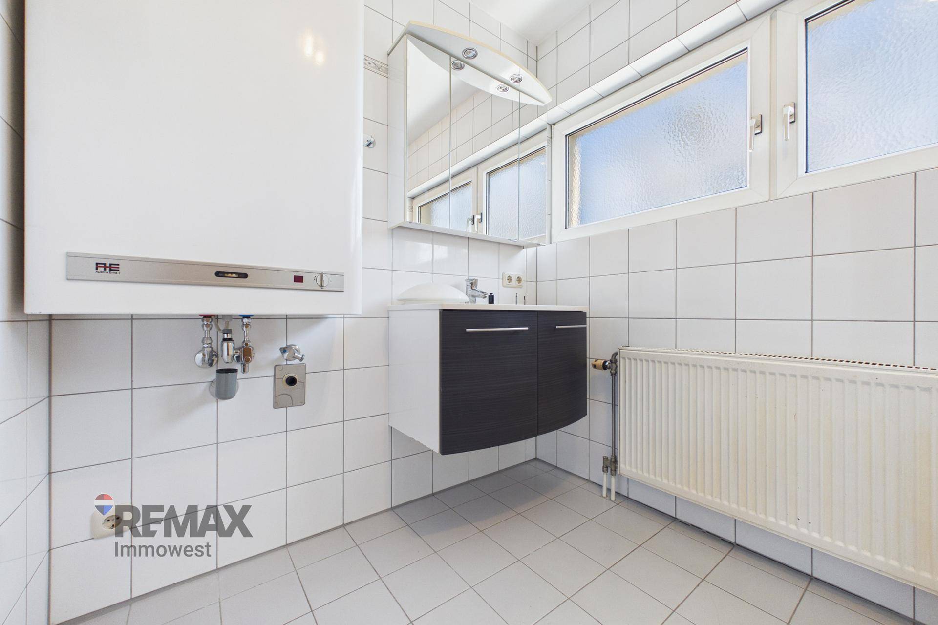 Gefliestes Badezimmer mit Waschbecken, Spiegelschrank und Warmwasserboiler, hell durch Fenster.