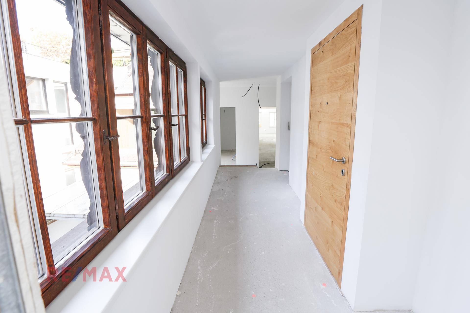 Langer Flur in einem Rohbau mit großen Holzfenstern und unfertigem Betonboden, bereit für den Ausbau.