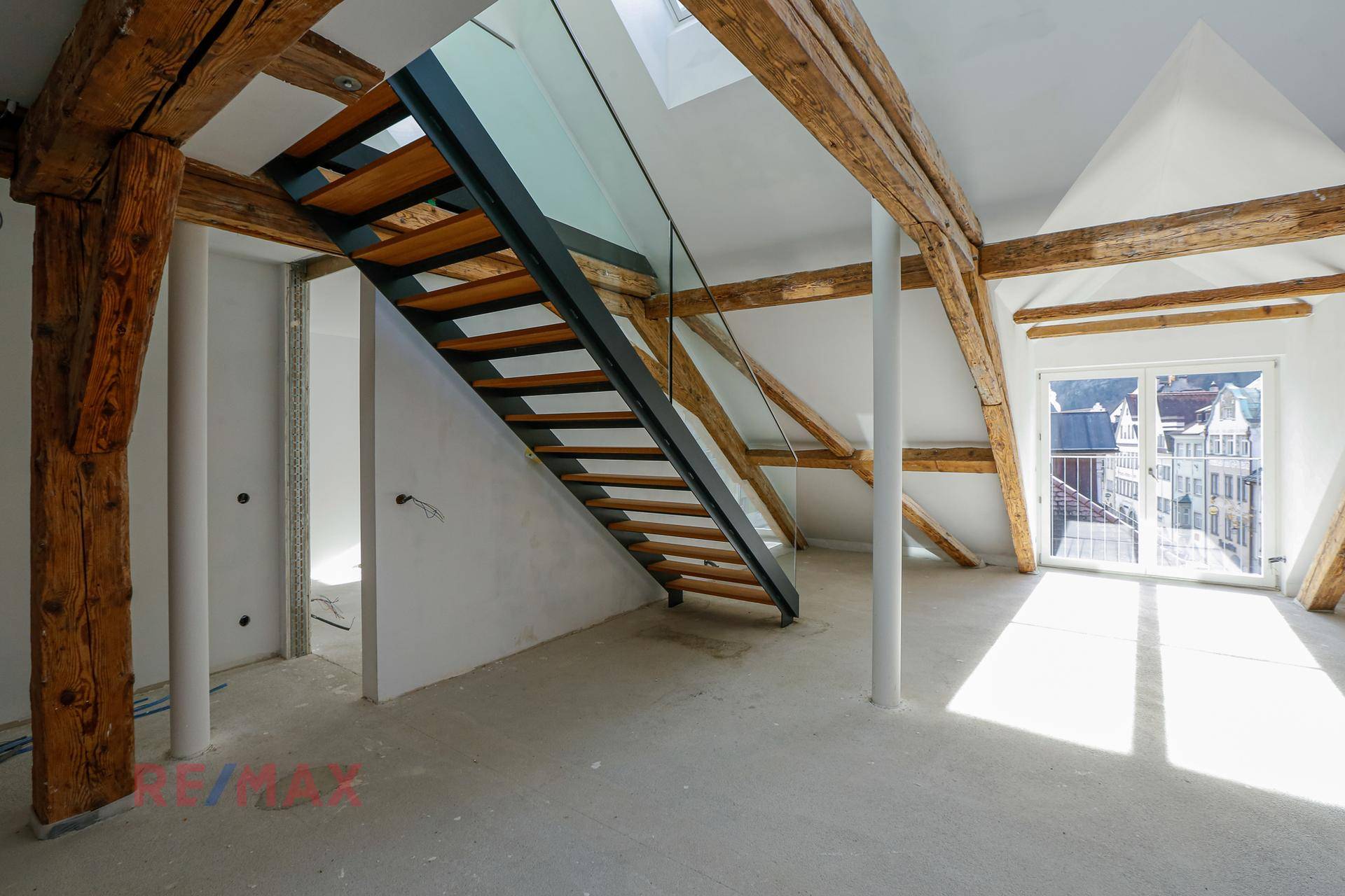 Offener Dachbodenbereich mit moderner Treppe aus Holz und Glas sowie freiliegenden Holzbalken und Tageslicht.