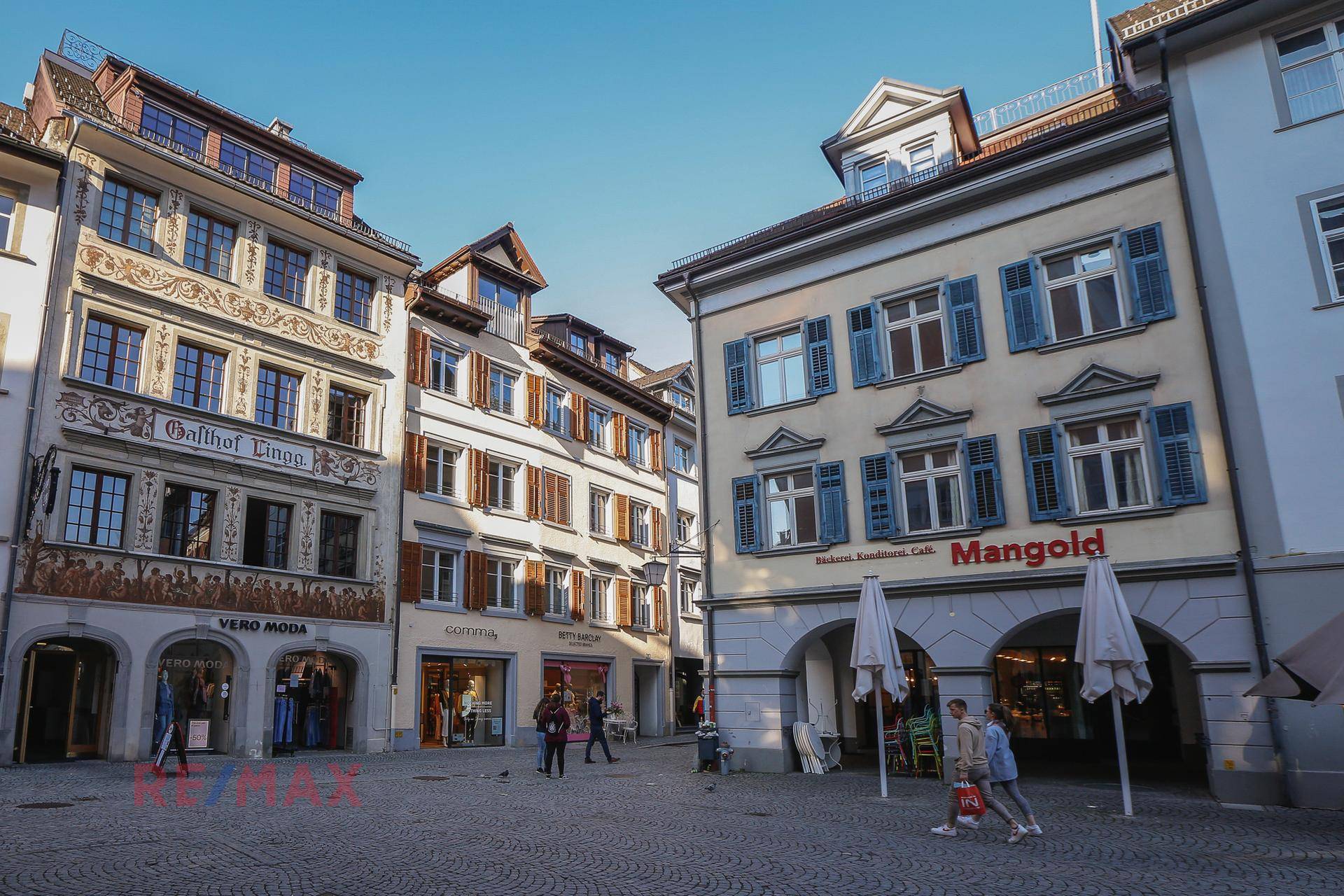 Historische Gebäude in einer belebten Gasse mit Geschäften und Restaurants im Stadtzentrum unter blauem Himmel.