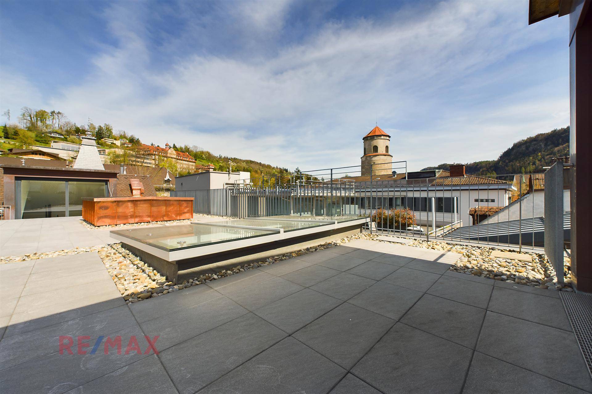 Große Dachterrasse mit Fliesenboden, Kiesbeeten und Blick auf die umliegende Stadt und grüne Hügel.