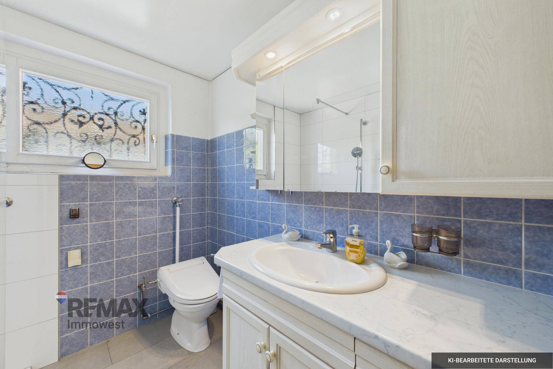 Badezimmer mit blauen und weißen Fliesen, Einzelwaschbecken, WC und Spiegelschrank, mit Fenster.