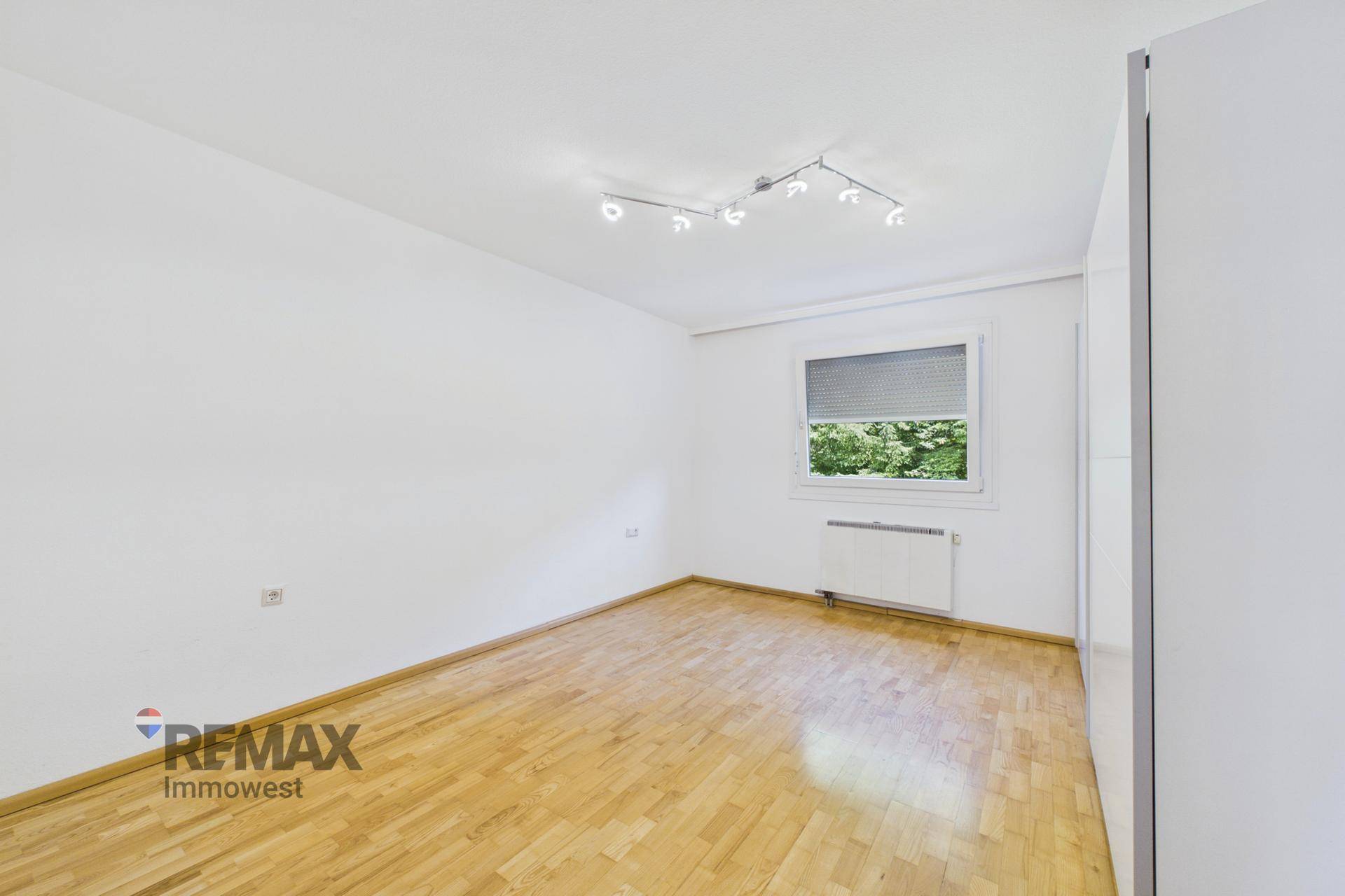 Helles Schlafzimmer mit Parkettboden, weiß gestrichenen Wänden und einem Fenster mit Blick ins Grüne.
