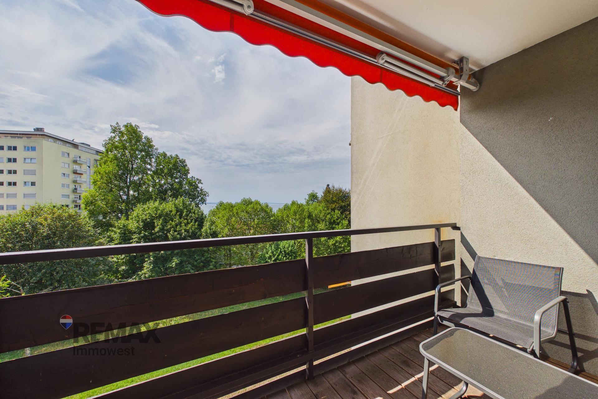 Möblierter Balkon mit Holzboden, roter Markise und Blick auf grüne Bäume.