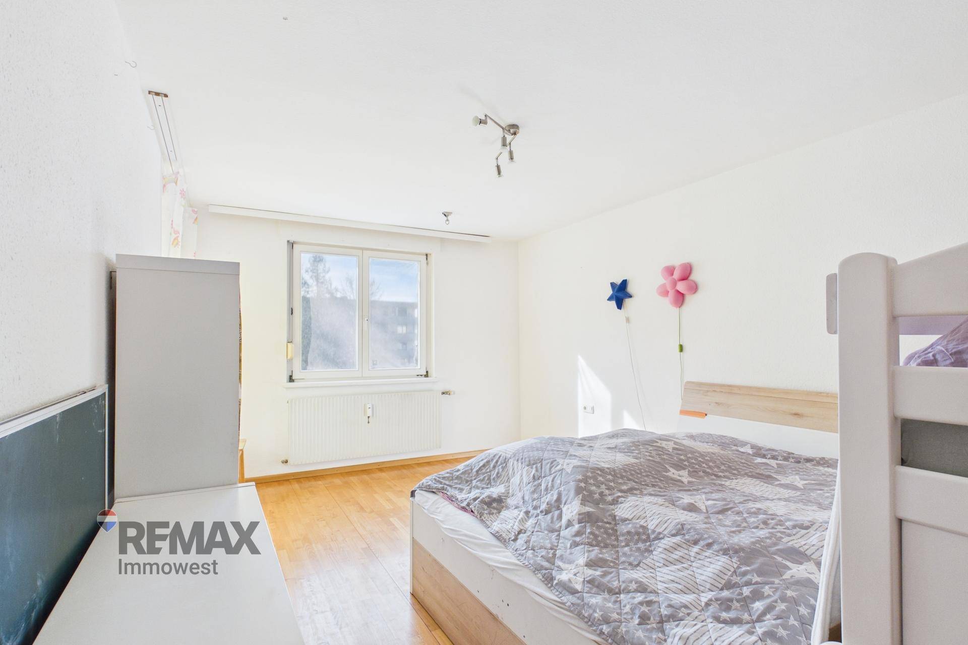 Helles Schlafzimmer mit großem Fenster, Holzboden und einfacher Einrichtung.