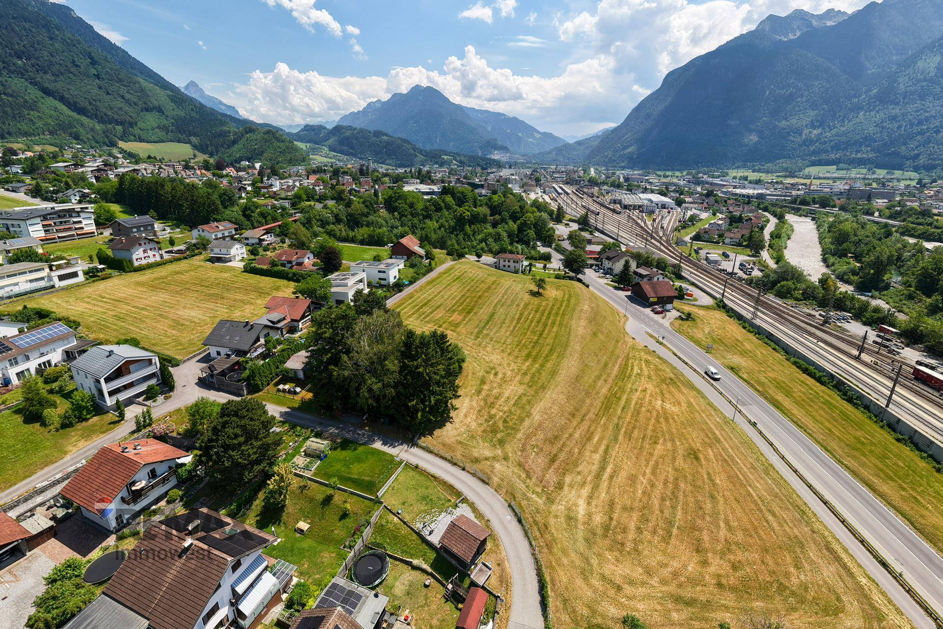 Panoramablick auf ein großes Feld, umgeben von Wohnhäusern, Straßen und Bahngleisen vor Bergpanorama.