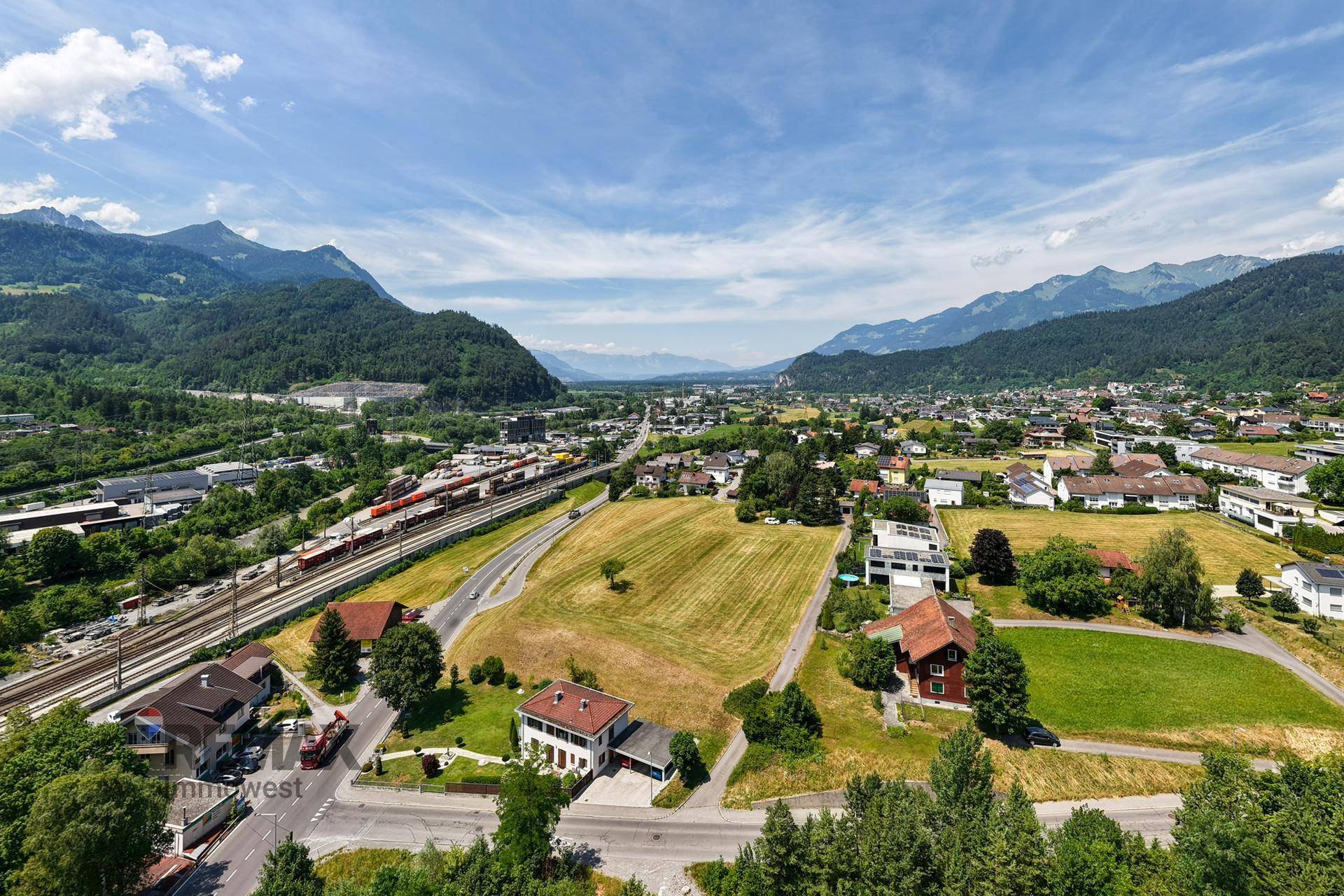 Luftaufnahme eines Feldes mit umliegenden Häusern, einer Straße und einer malerischen Berglandschaft.