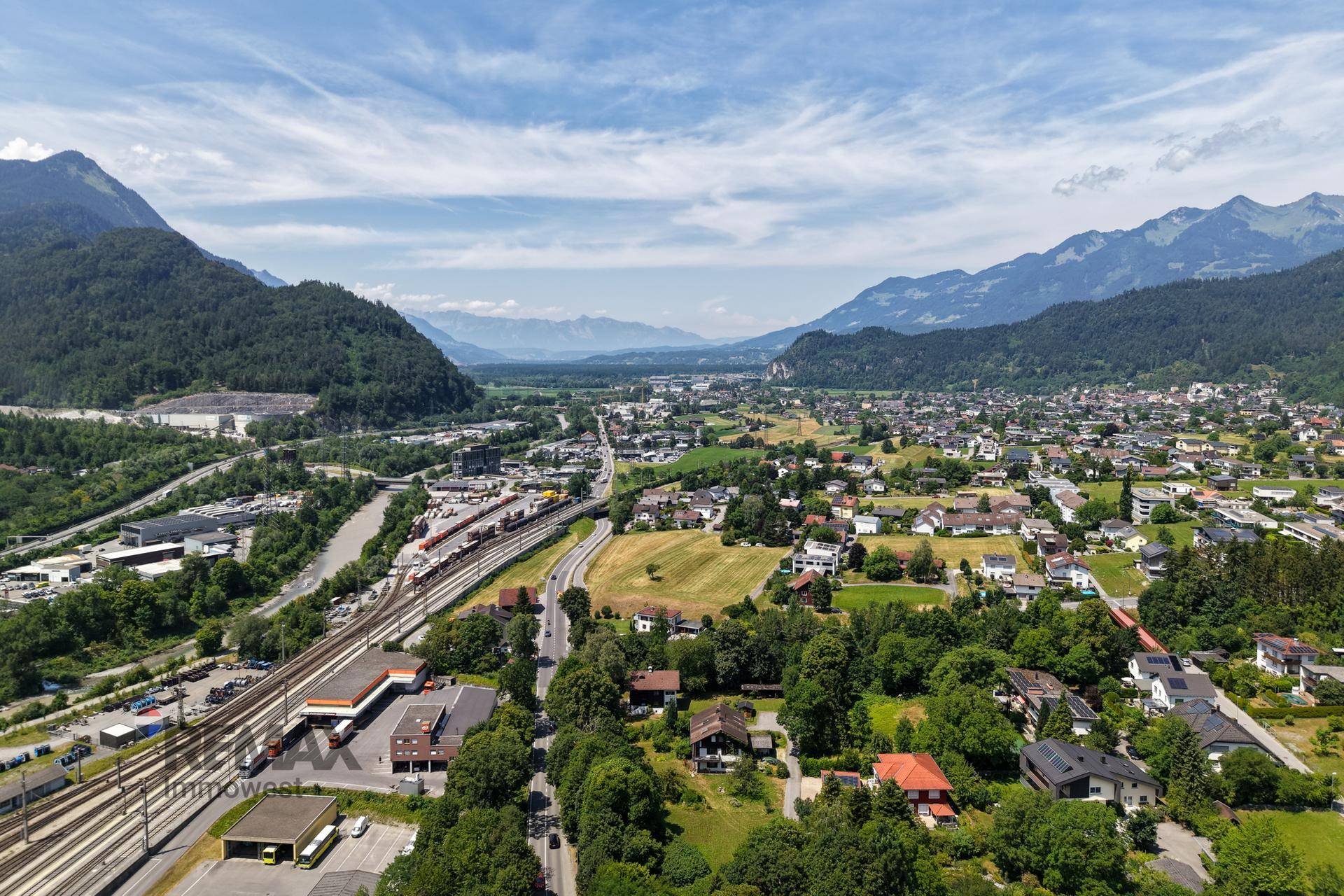 Weitläufige Luftaufnahme eines Tals mit einer Siedlung, Bahngleisen und Bergen am Horizont.