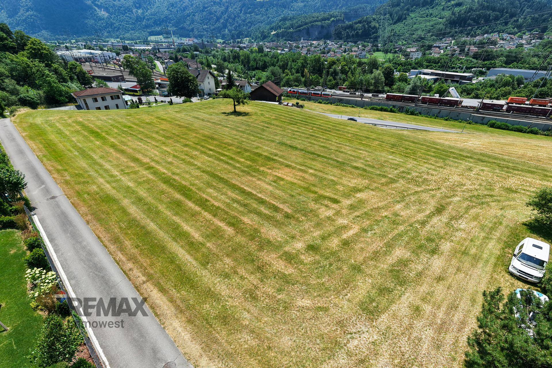 Luftaufnahme eines großen Feldes neben einer Straße mit einem Dorf und Bahngleisen in der Ferne.