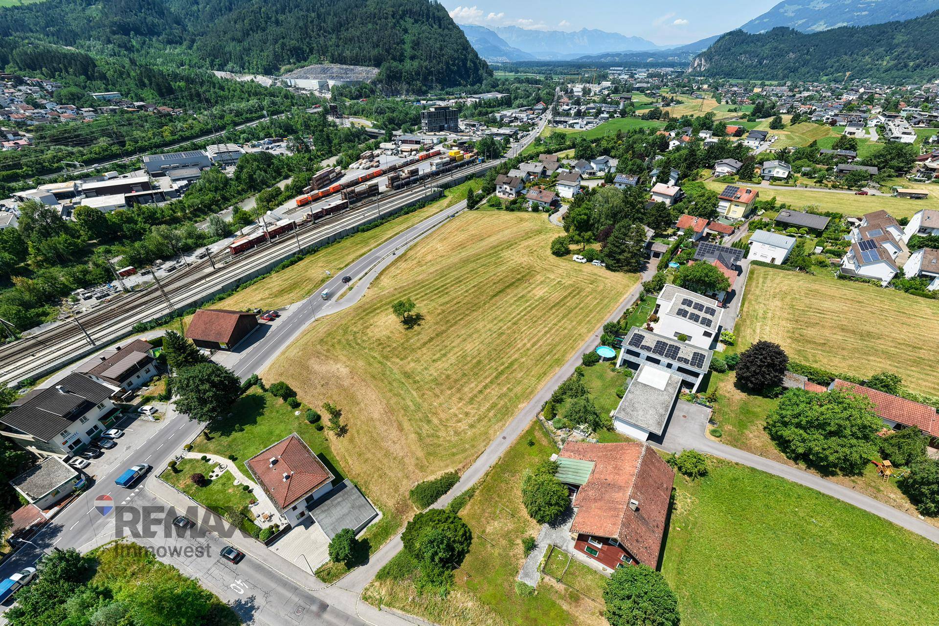 Luftaufnahme von Feldern, Häusern und Straßen mit Bahngleisen und Bergen im Hintergrund.