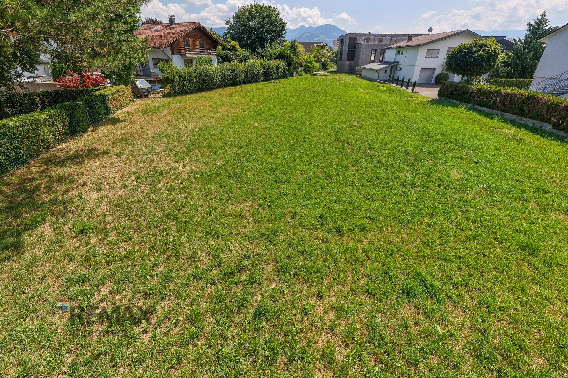 Weitläufiges, sonniges Grundstück mit Rasen und Hecken, umgeben von Wohnhäusern und Bergen im Hintergrund.