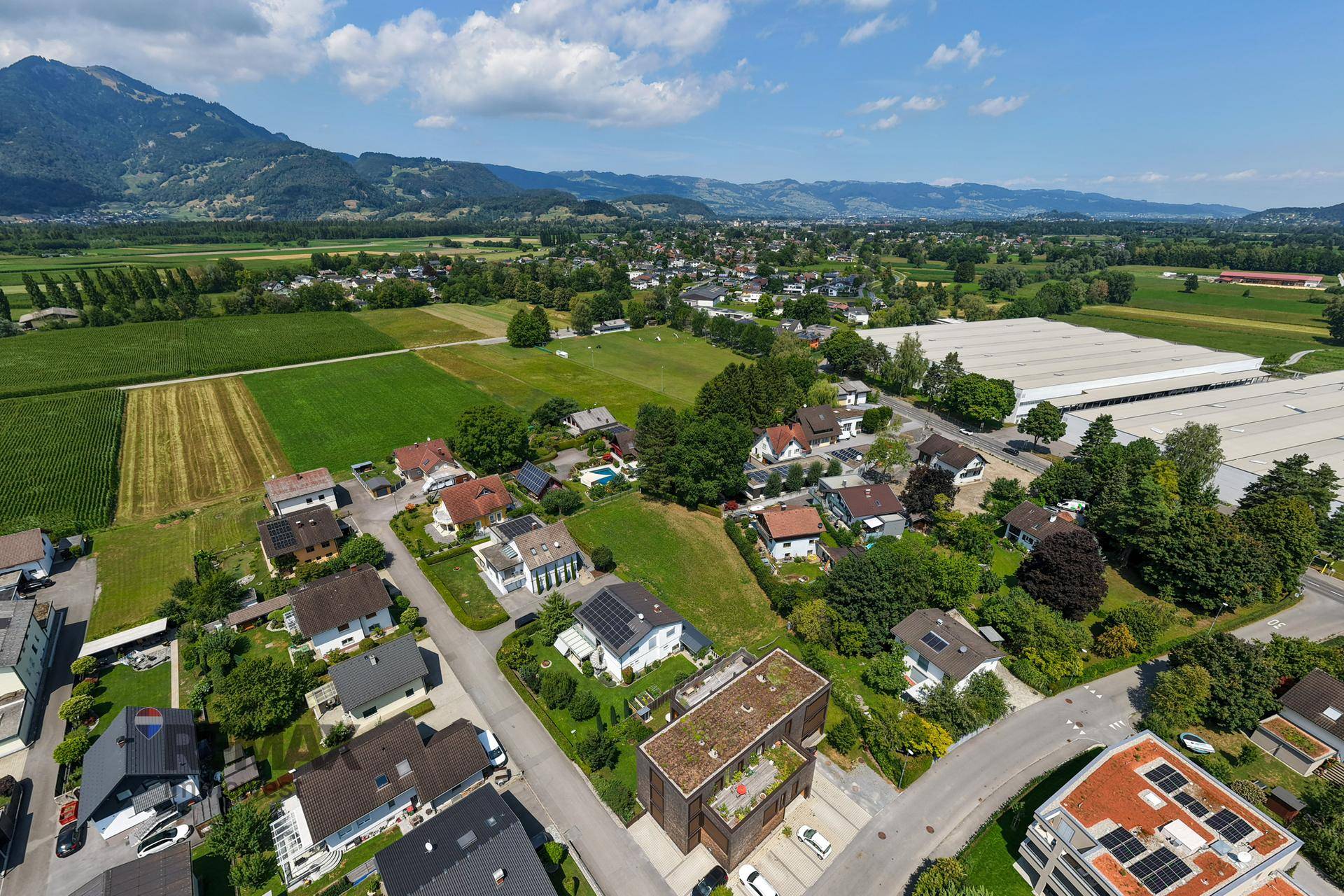 Luftaufnahme einer Wohnsiedlung mit grünen Feldern und Bergen im Hintergrund, zeigt die Umgebung des Grundstücks.