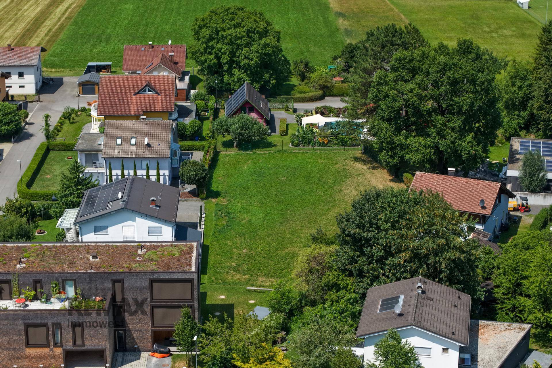 Luftaufnahme des Grundstücks in einer grünen Umgebung, umgeben von Häusern, Feldern und Bergen im Hintergrund.