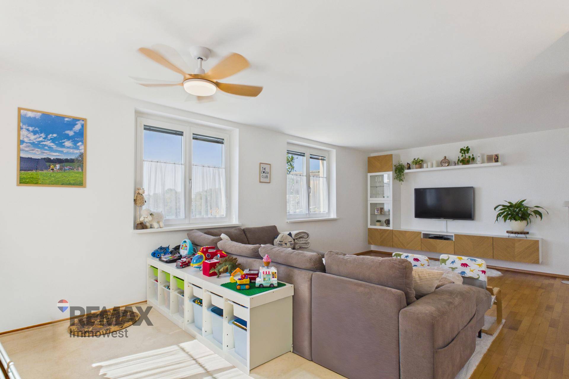 Helles Wohnzimmer mit Spielbereich, Deckenventilator und Blick aus den Fenstern.