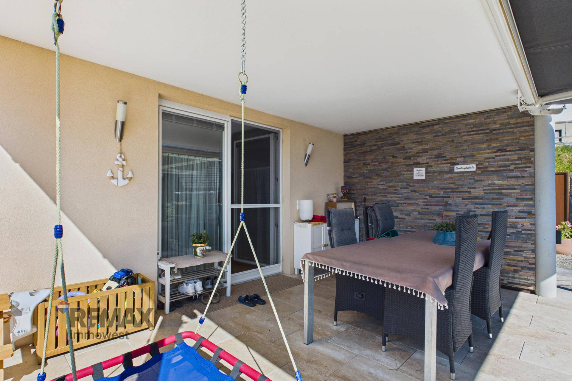 Überdachte Terrasse mit Esstisch, Steinwand und Schaukel für entspannte Stunden im Freien.