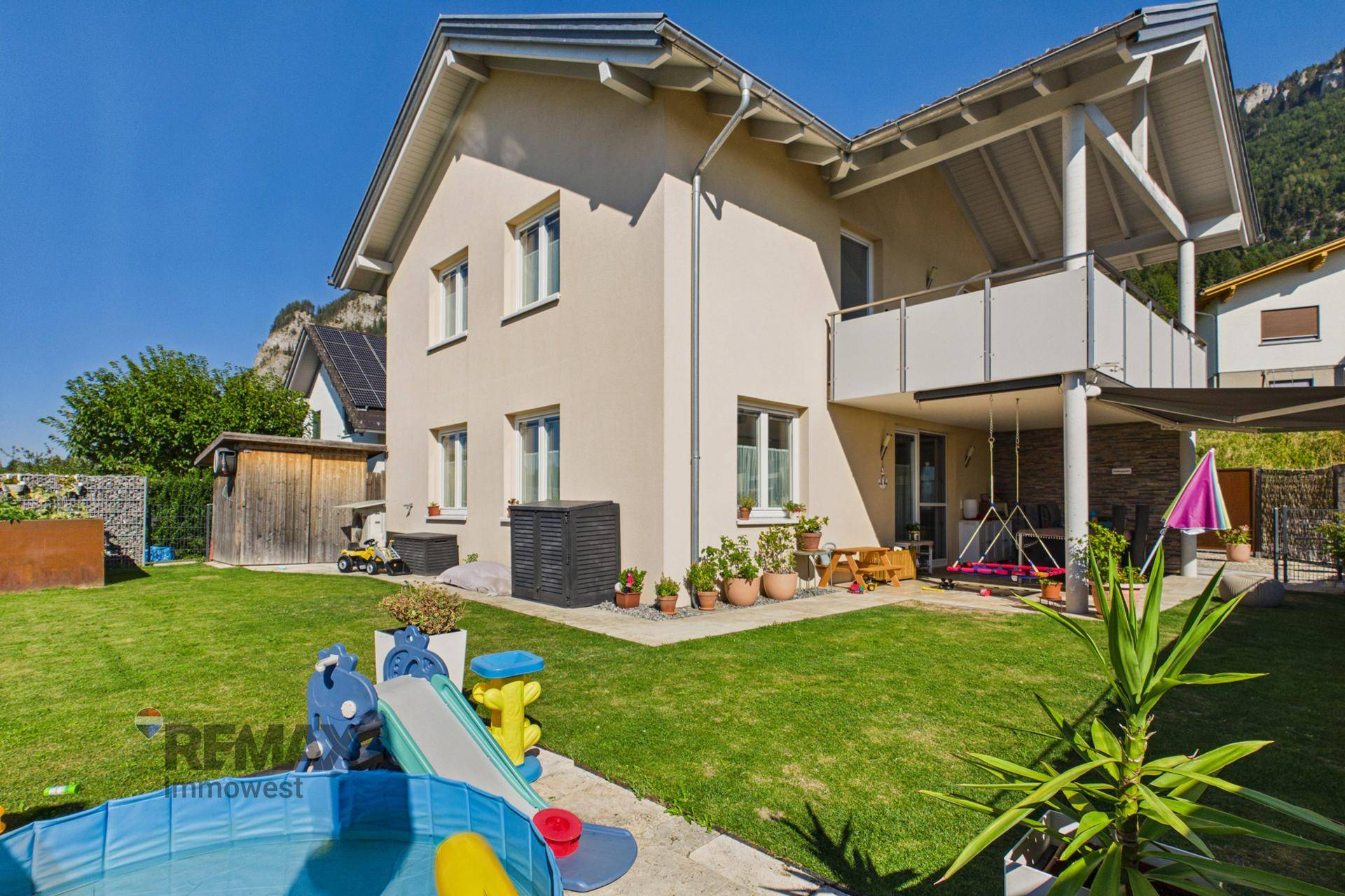 Ansicht des Hauses von der Gartenseite mit Rasenfläche, Spielgeräten und überdachter Terrasse.