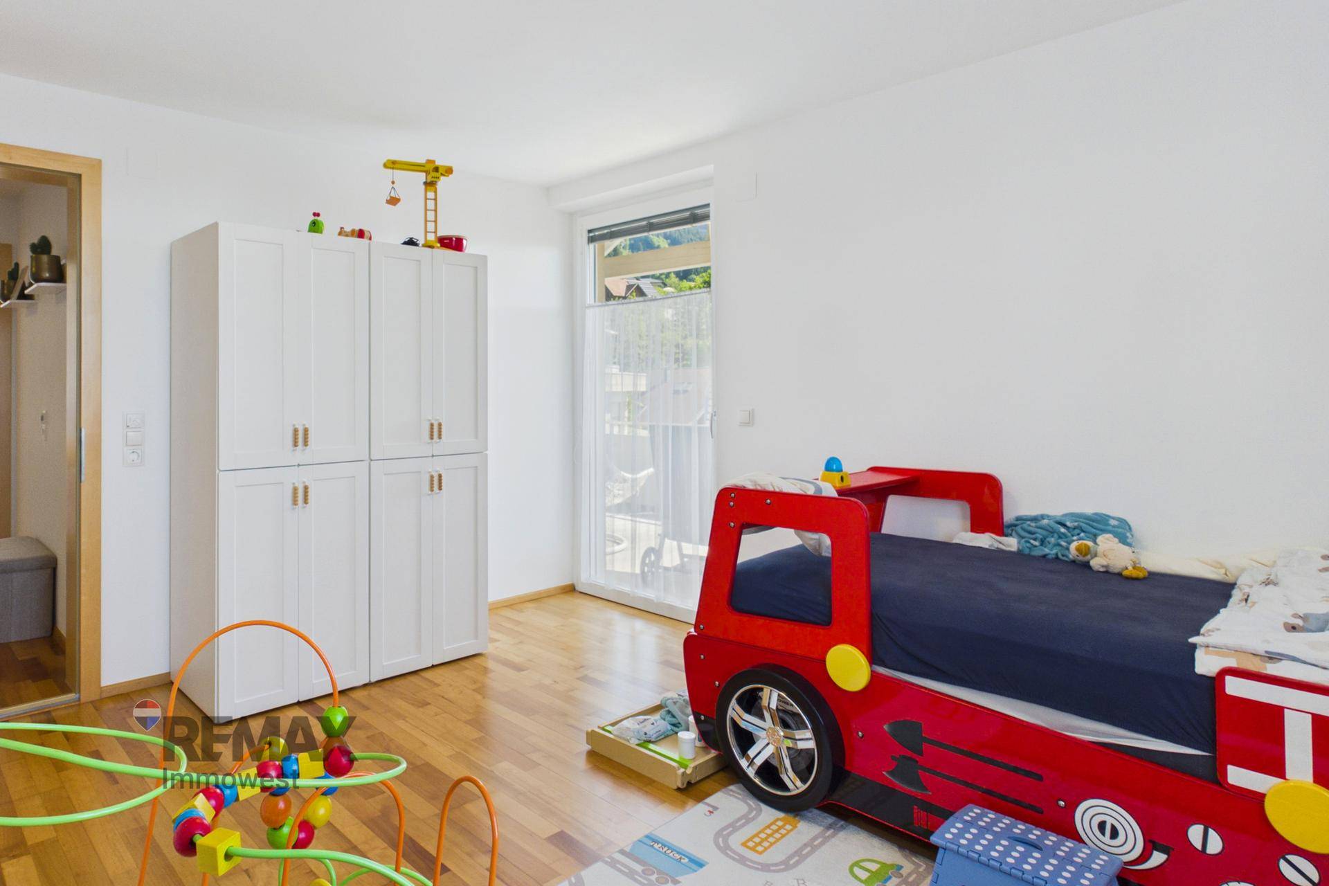 Kinderzimmer mit Spielzeug, Schrank und Zugang zu einem kleinen Balkon oder Terrasse.