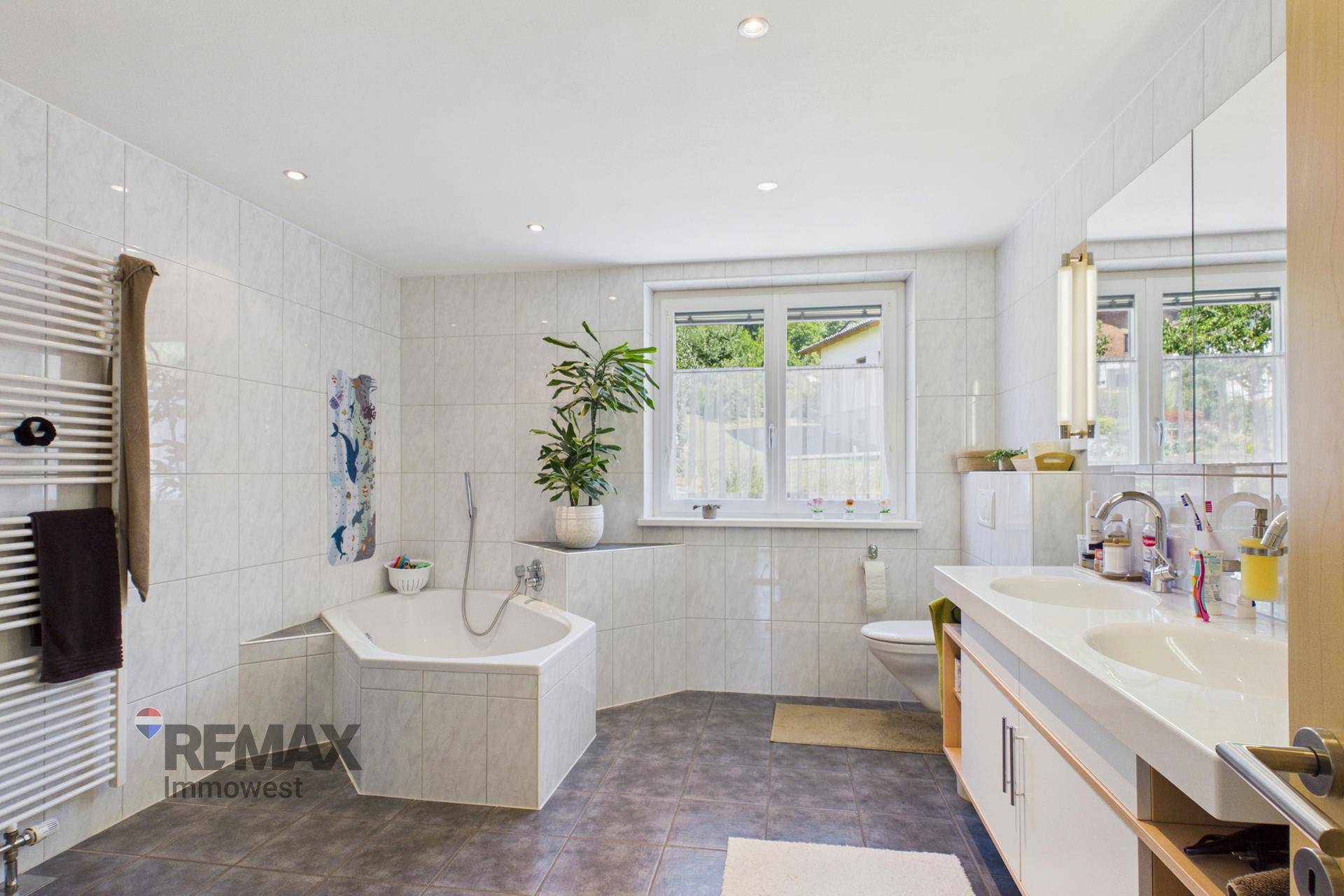 Helles Badezimmer mit Eckbadewanne, Doppelwaschbecken, Handtuchheizkörper und großem Fenster.