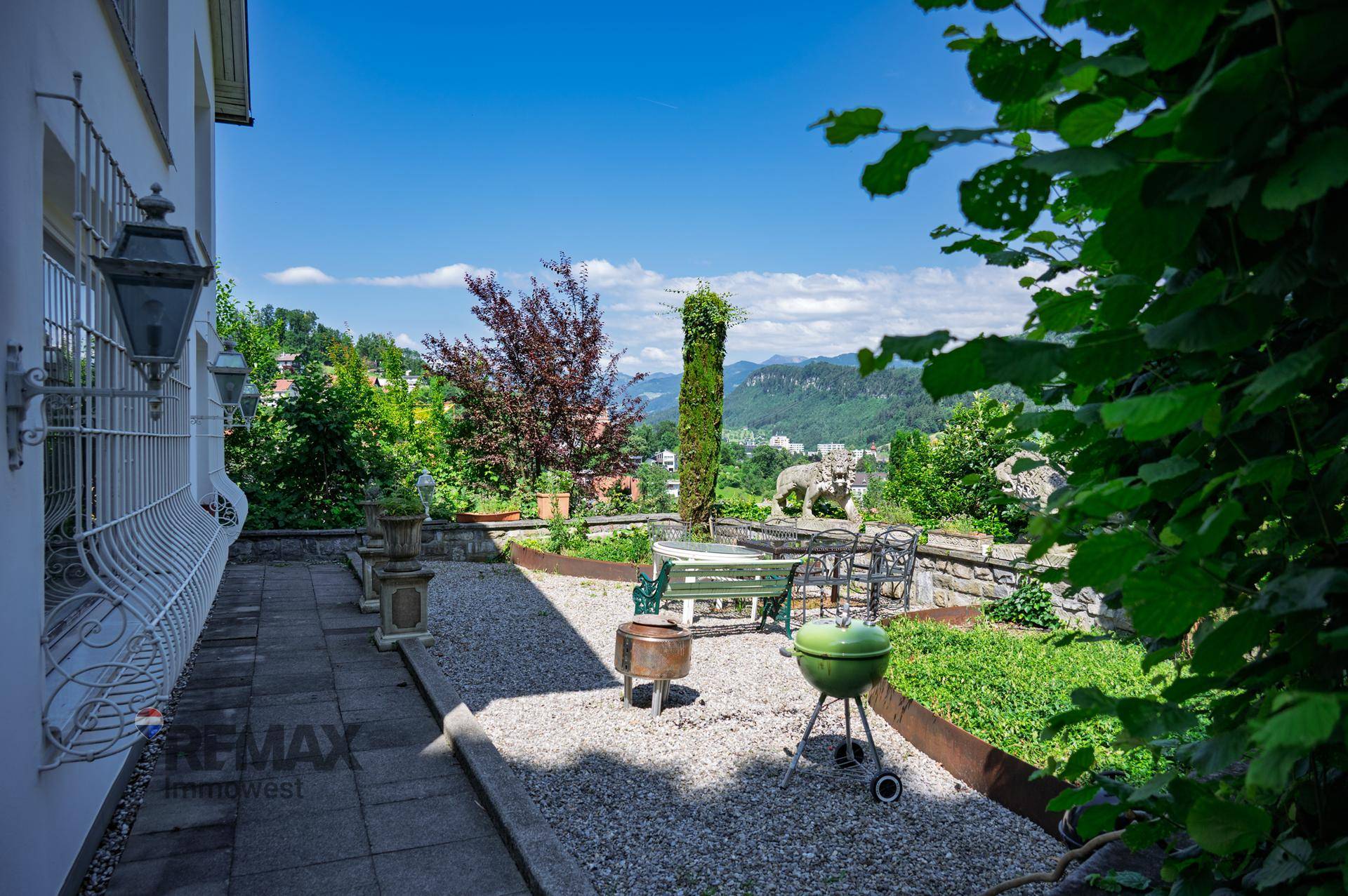 Sonniger Terrassenbereich mit Kiesboden, Gartenmöbeln und Grill, umgeben von Pflanzen und Weitblick.