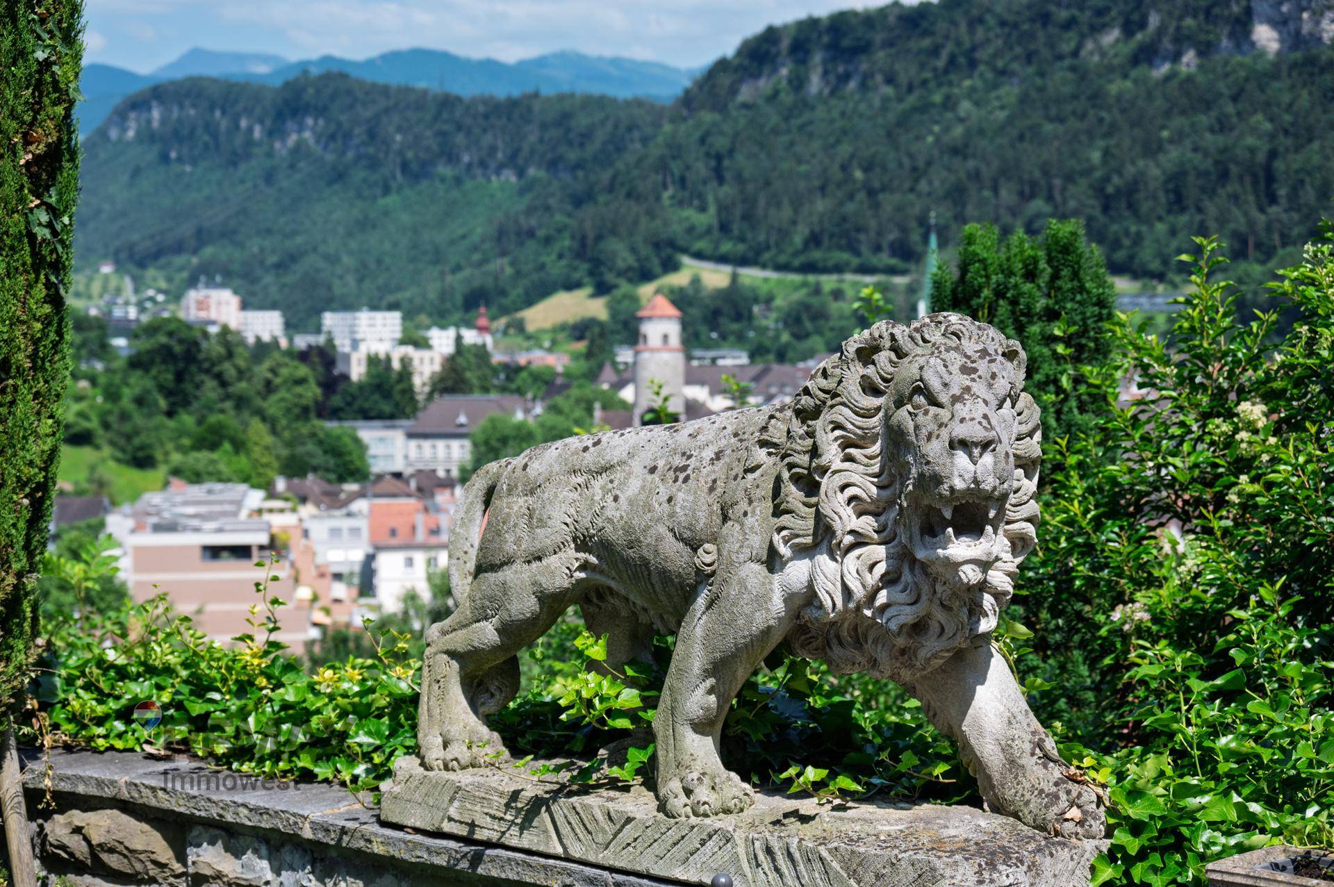 Löwenstatue im Vordergrund mit Panoramablick auf die Stadt und die bewaldeten Berge im Hintergrund.