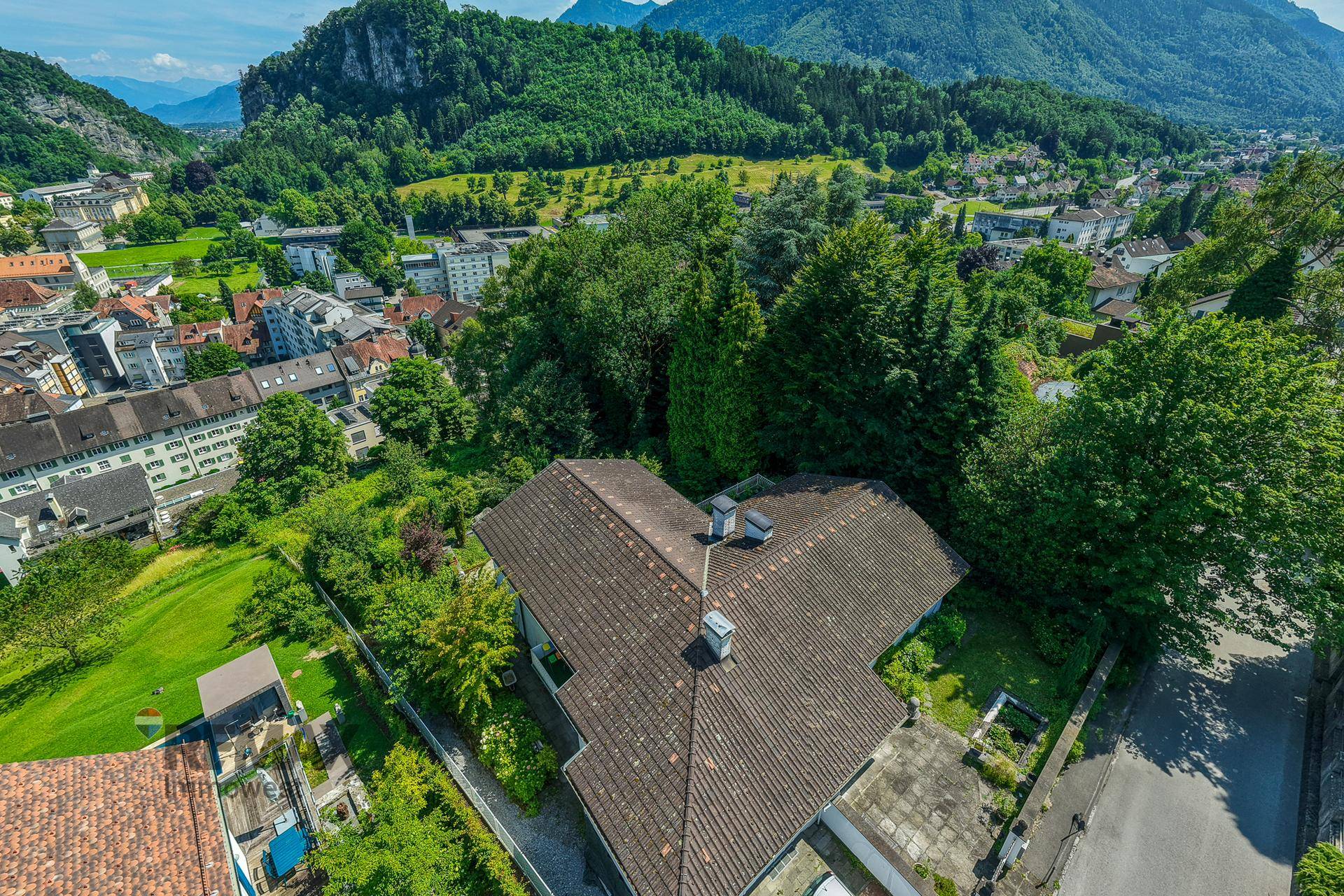 Luftaufnahme eines Hauses mit Satteldach, umgeben von viel Grün und beeindruckendem Bergblick im Hintergrund.